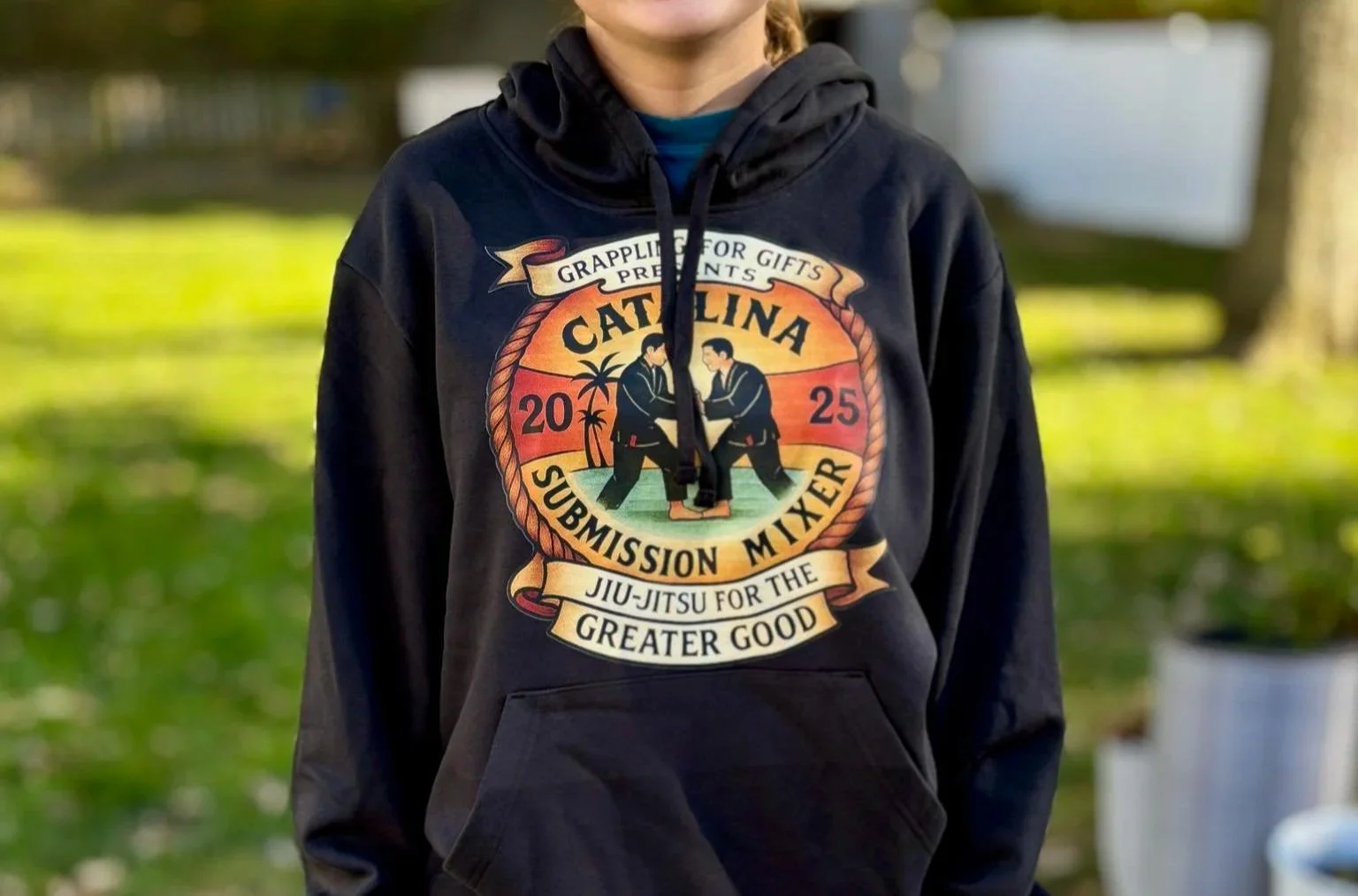 Person wearing a black hoodie with a graphic design for a submission mixer event, featuring two people practicing Jiu-Jitsu, palms, and text about the event held in 2025 in Carolina.