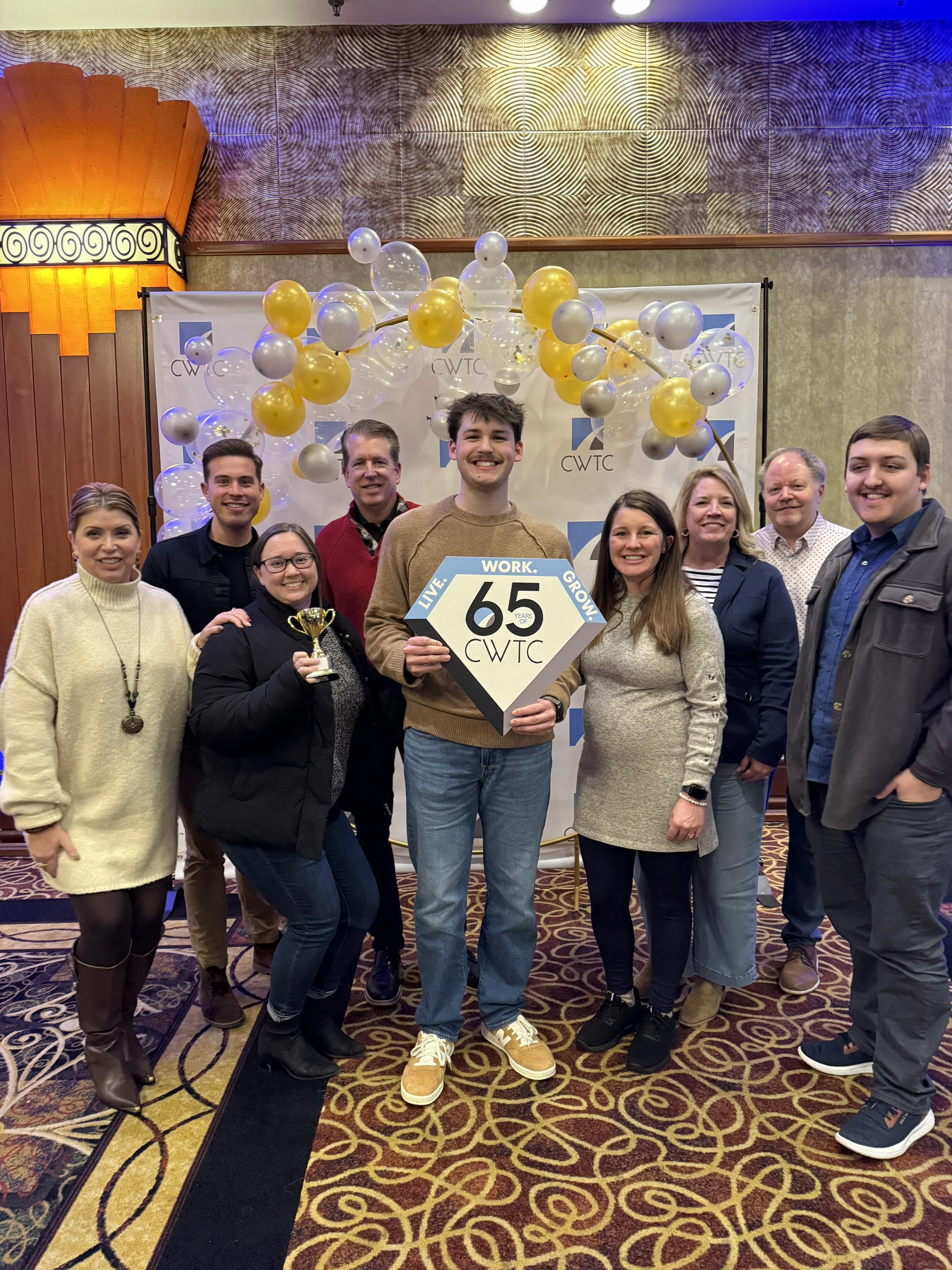 Group of nine people celebrating at an event, with a man holding a sign that reads '65 CWTC' and a small trophy, standing in front of a decorative backdrop with balloons and the CWTC logo.