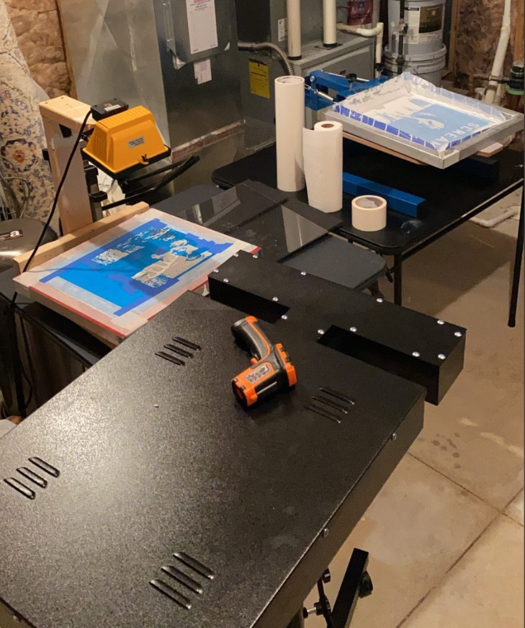 A workshop workspace with black metal equipment, a cordless drill on top, paper towels, packaging materials, and various tools and supplies.