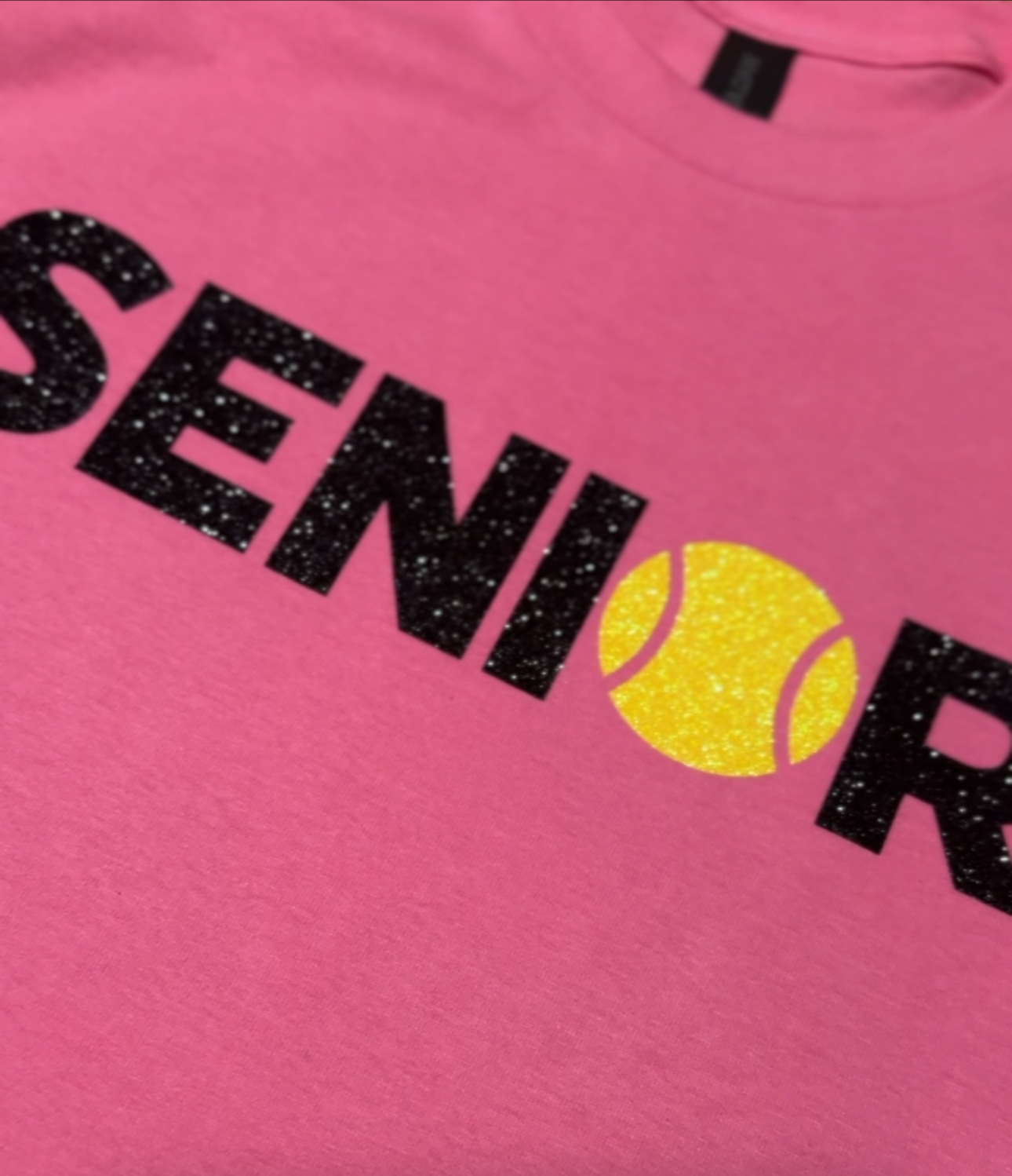 Close-up of a pink T-shirt with black glittery text and a yellow glittery tennis ball graphic.