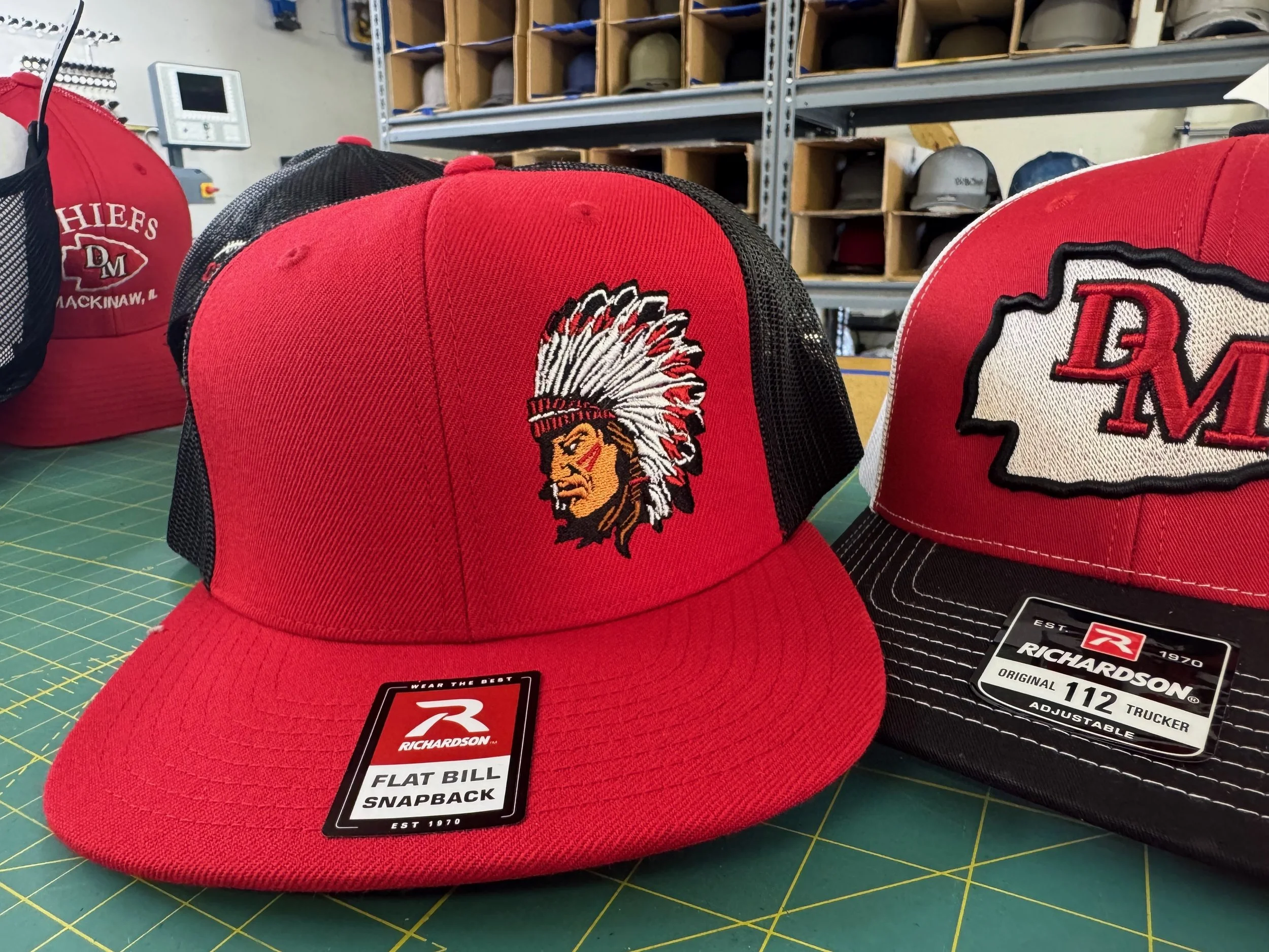Red and black trucker caps with embroidered team logos and tags on a green cutting mat, with a background of shelves with helmets and other items.