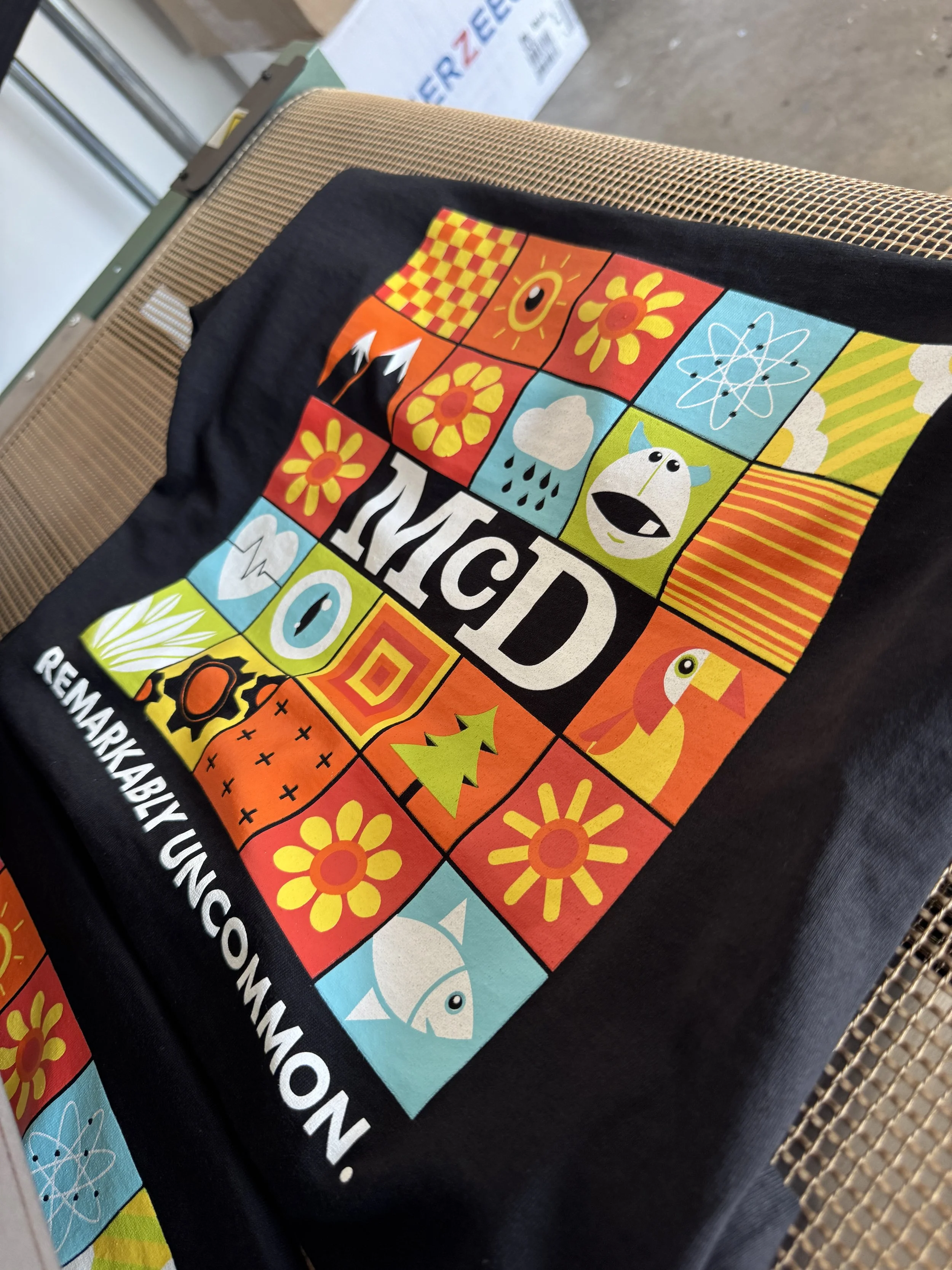 A black cloth with colorful squares featuring various illustrations including flowers, animals, weather, and scientific symbols, with the text 'MCDC' and 'Remarkably Uncommon'.