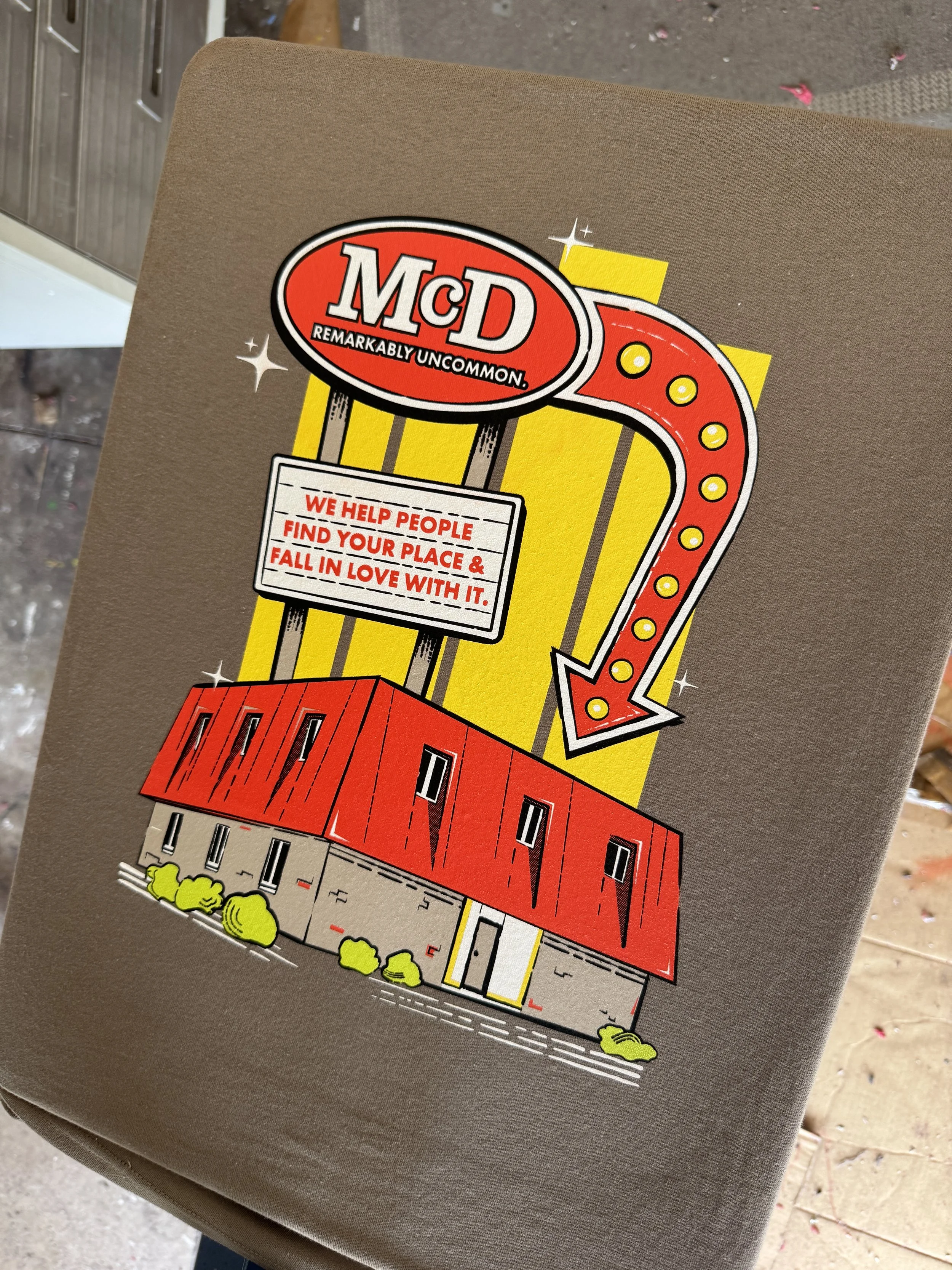 A vintage-style graphic illustration of a building with a large, neon-like sign that reads "McD" with the slogan "Remarkably Uncommon" underneath. A large arrow points downward to the building, which has a red roof and is surrounded by small green bu