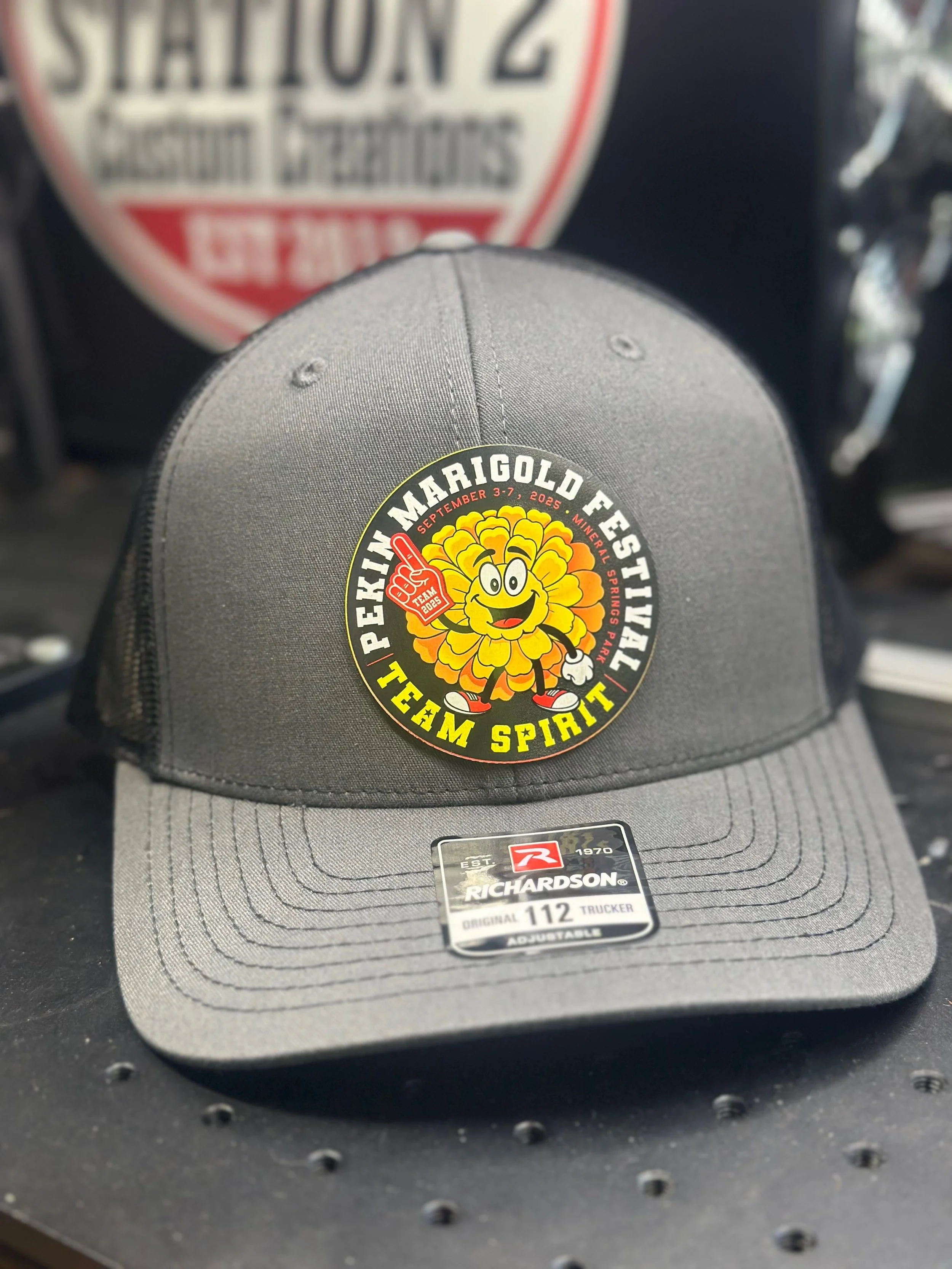 A gray baseball cap with a colorful patch on the front that reads "Pekin Marigold Festival Team Spirit" and features a cheerful marigold flower character holding a foam finger, with event details and dates. The cap is placed on a black surface with a blurred background.
