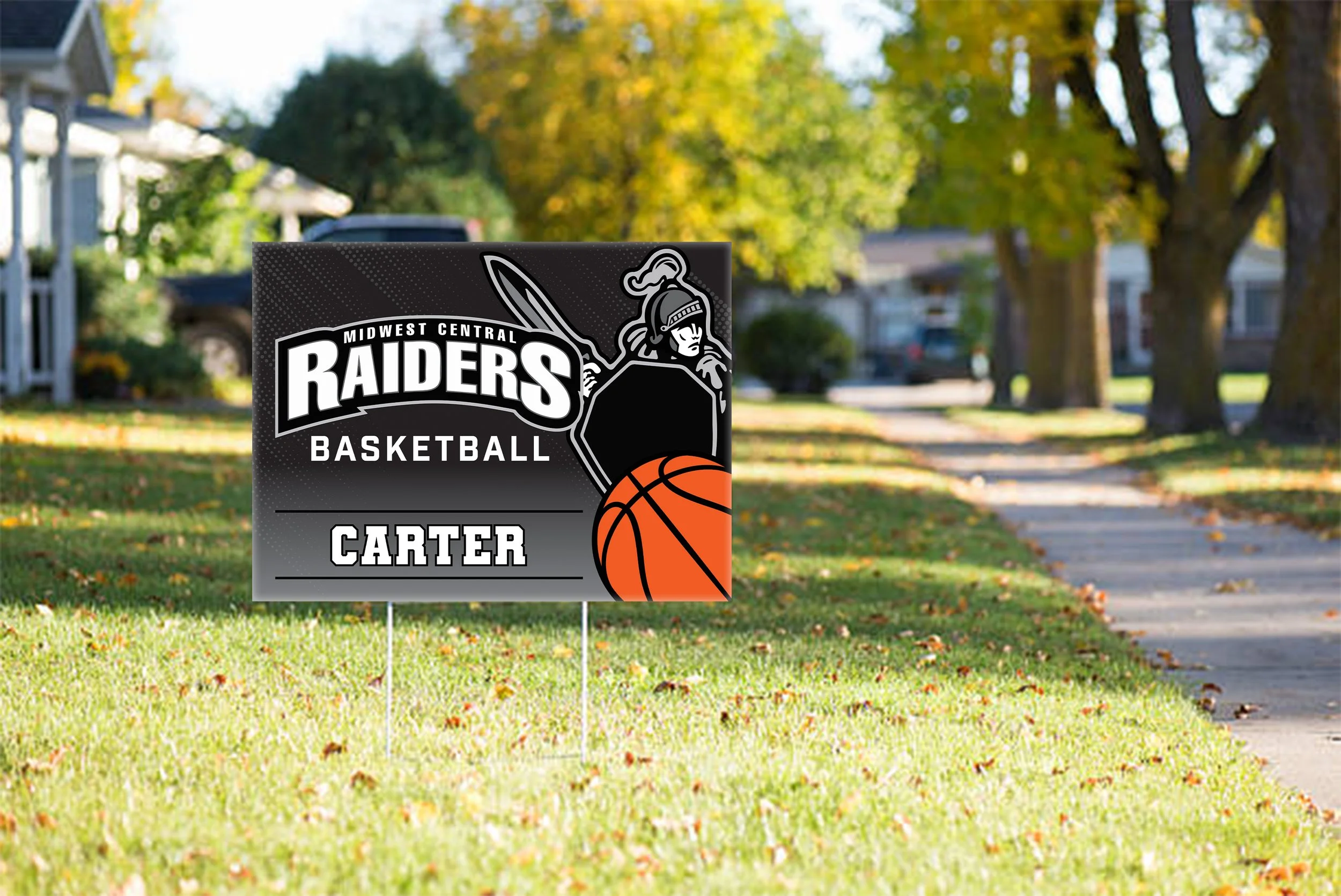 Yard sign for Carter with a Midland Central Raiders Basketball team theme, featuring a knight with a sword and shield, and a basketball graphic, on a grassy front lawn with trees and houses in the background.