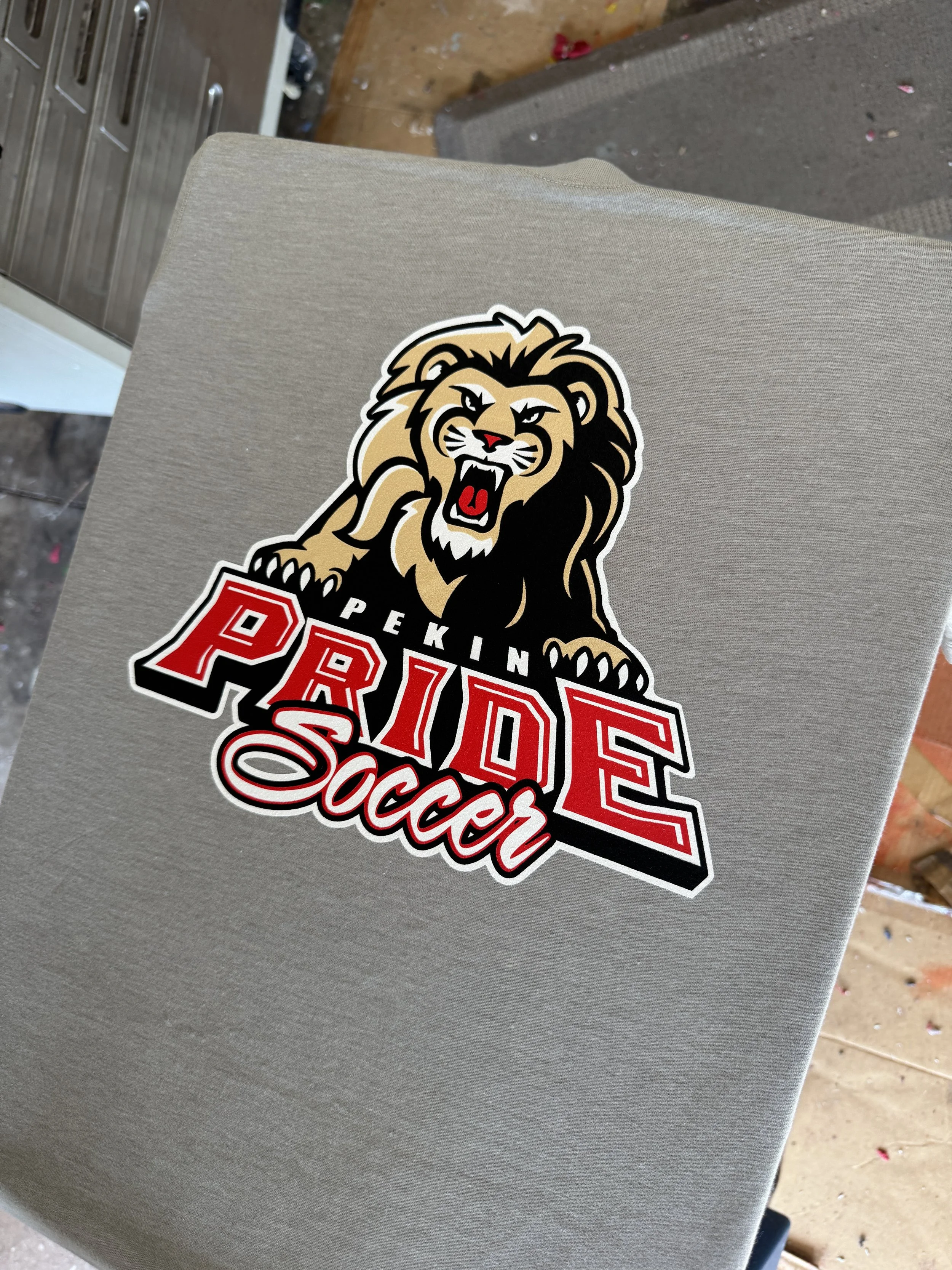 A sticker of a fierce lion with a mane, open mouth showing teeth, and raising one paw, with the words "PEKING PRIDE Soccer" underneath, attached to a light-colored surface.