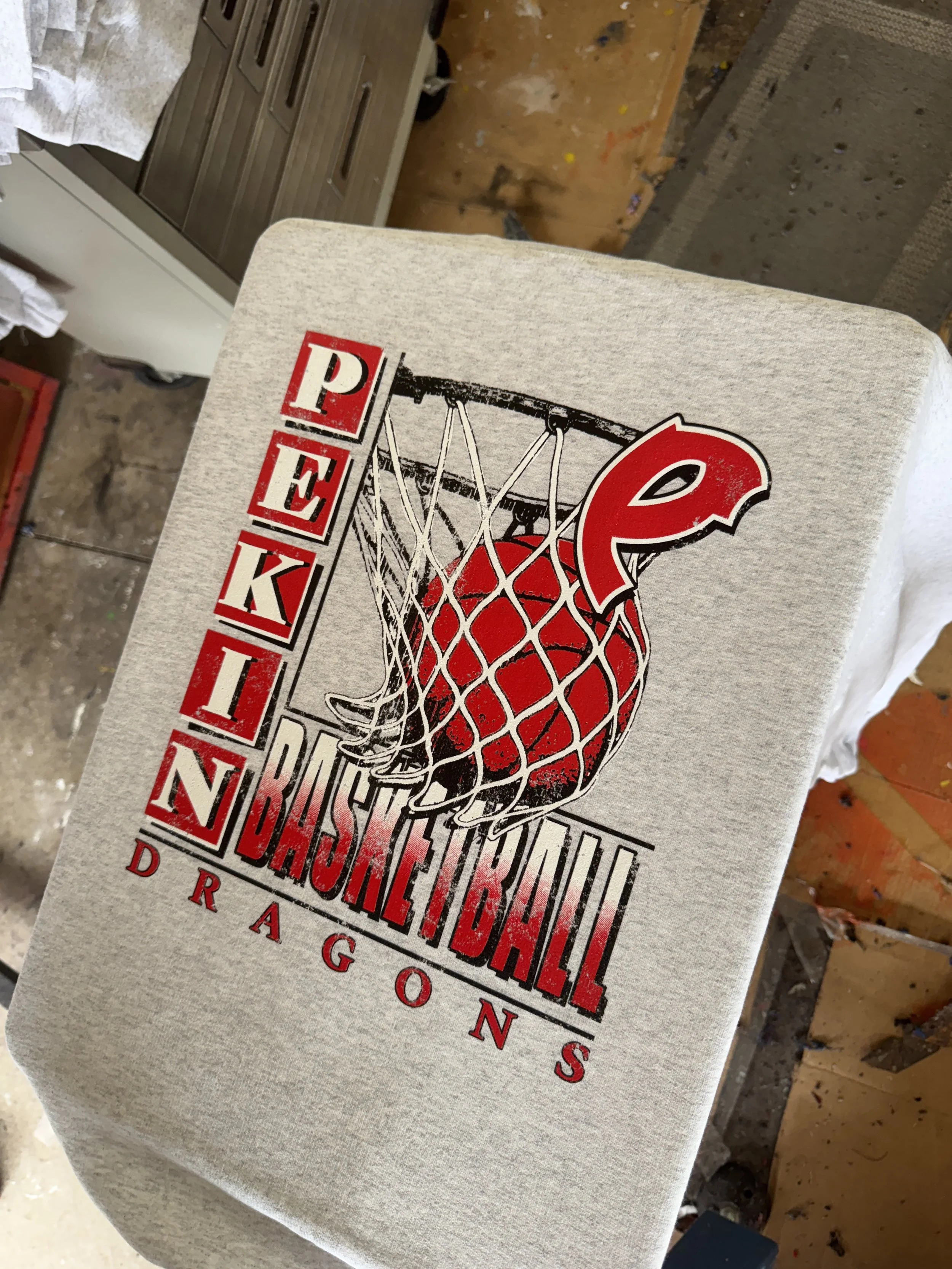 Design of a basketball going through a hoop, with text 'Pekin Baseball Dragons' in red and white lettering.