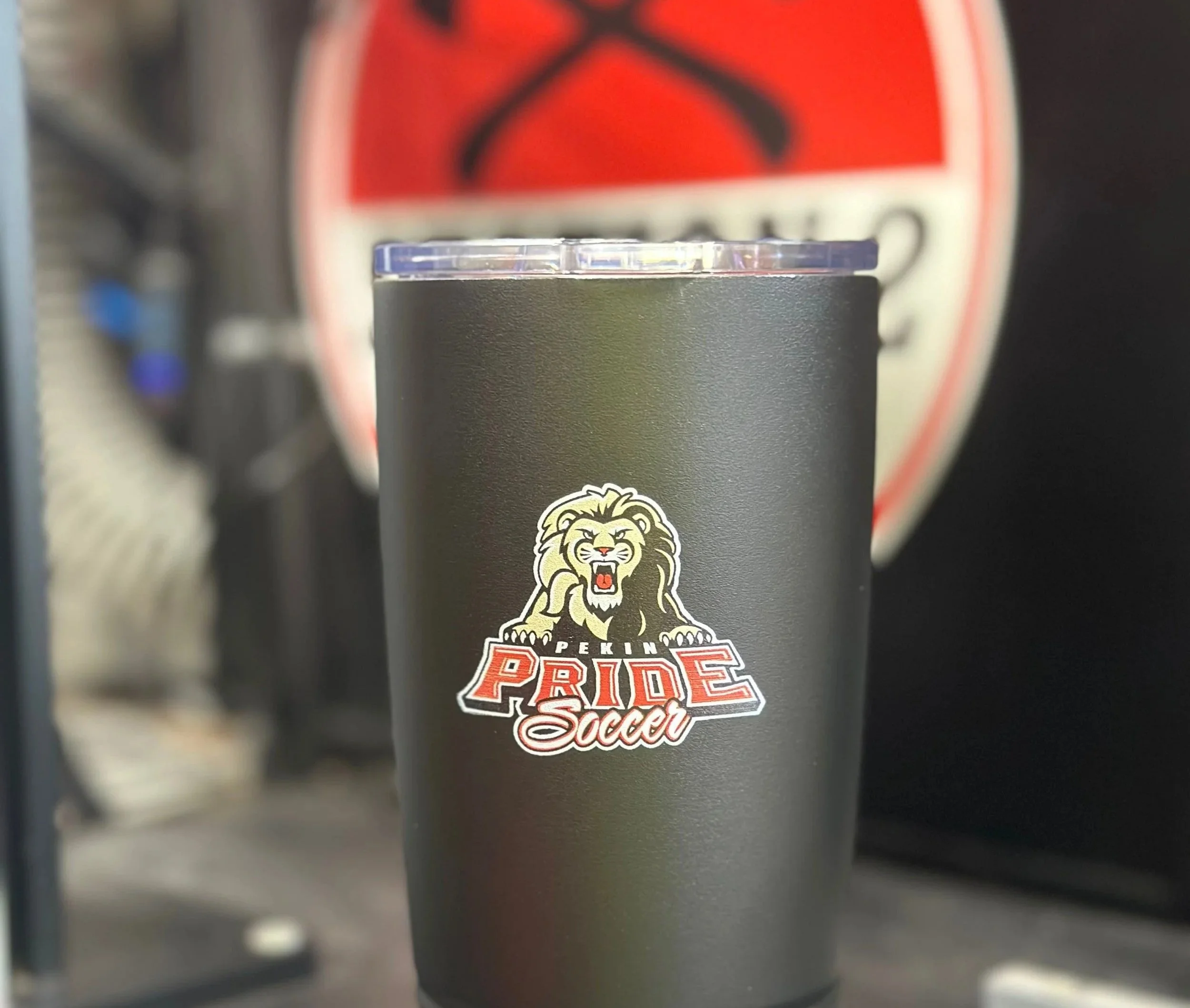 A black tumbler cup with a 'Pekin Pride Soccer' logo featuring a roaring lion, placed in front of a blurred background with a red and black caution sign.