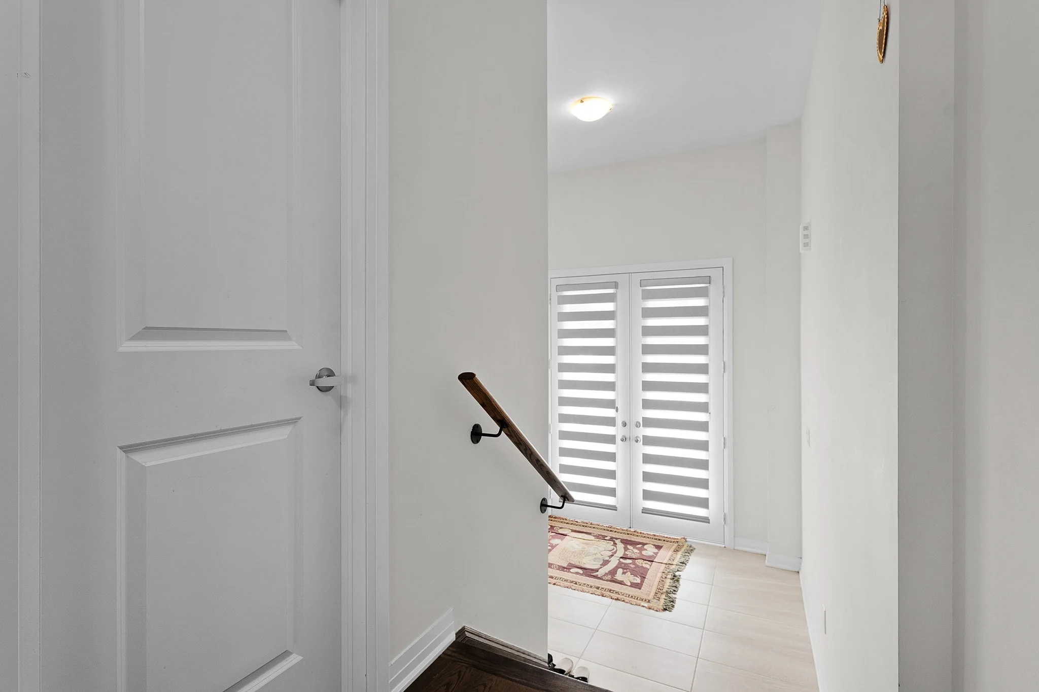 Interior view of a house hallway showing a white door on the left, a staircase with a wooden handrail, a window with white shutters, and a marble floor rug. The hallway walls are white, and there is a ceiling light fixture.