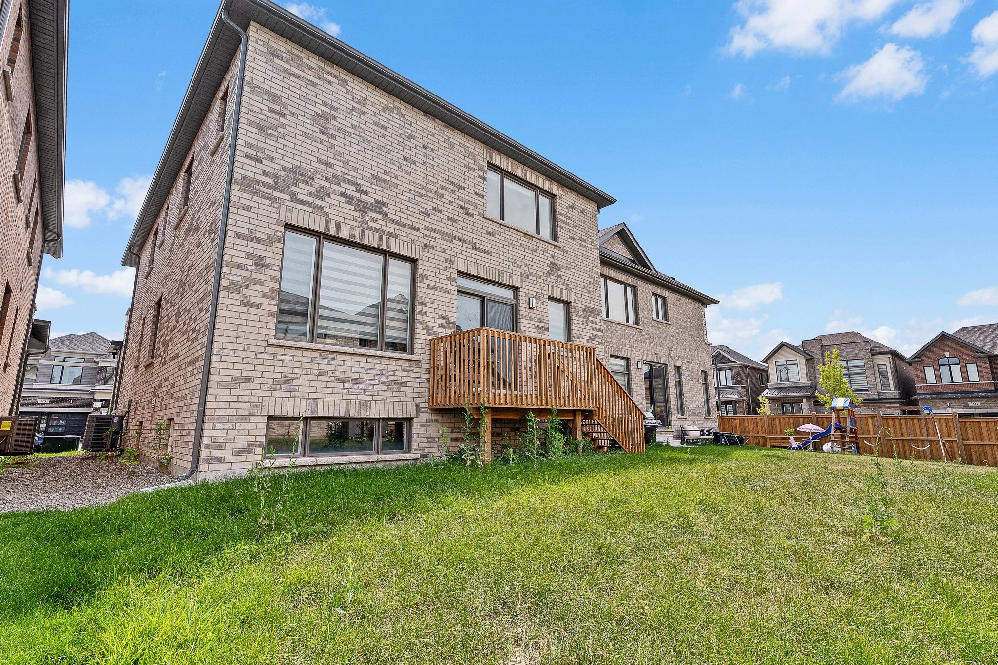 Backyard view of a multi-story brick house with a small wooden deck, surrounded by a fenced yard and neighboring houses under a partly cloudy sky.