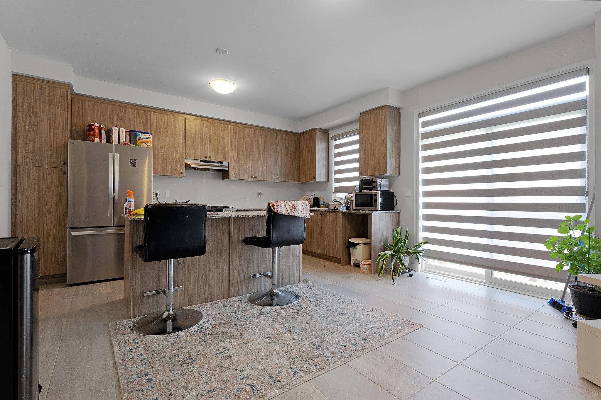 Modern kitchen with wooden cabinets, stainless steel refrigerator, microwave, black chairs at a kitchen island, large window with zebra blinds, potted plants, and a rug on tile flooring.