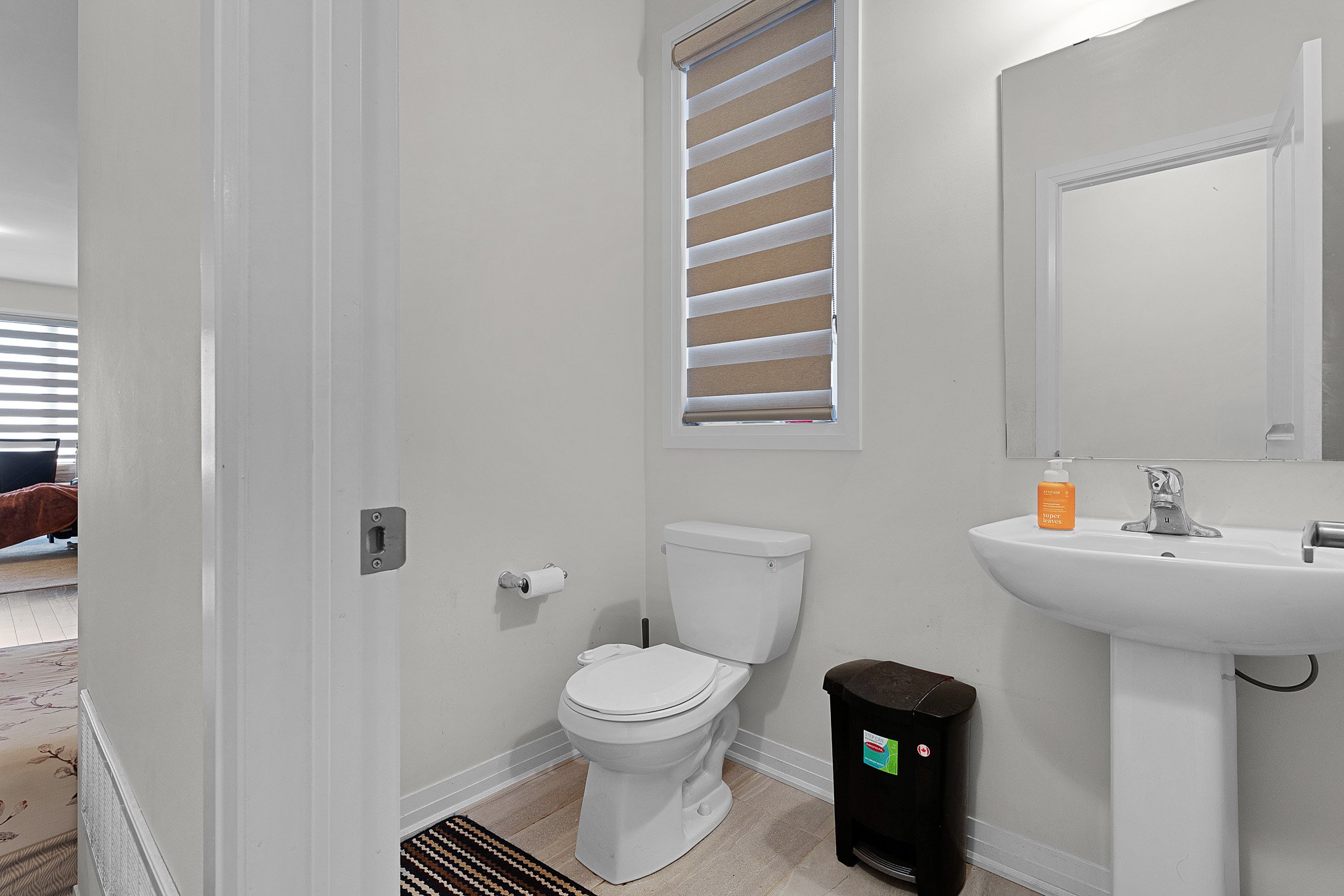 A small bathroom with a white toilet, a white sink with a mirror above it, a small black cabinet, a brown wastebasket, and a window with beige and white horizontal blinds. There is a bottle of hand soap on the sink.
