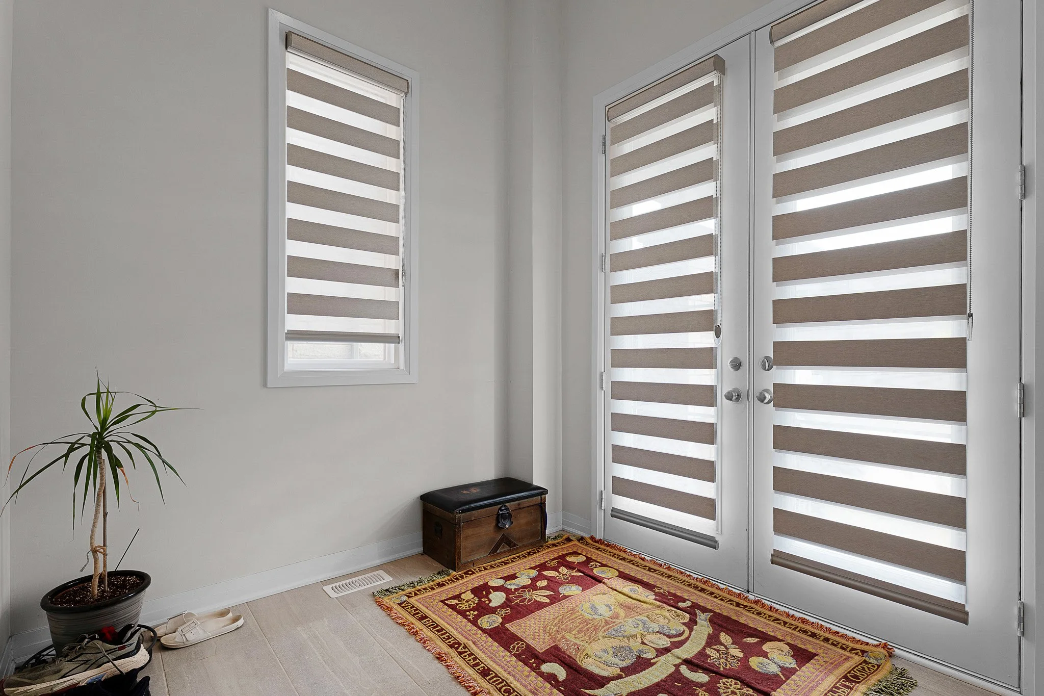 Entryway with a window and a door with striped shades, small potted plant, wooden chest, and a colorful patterned rug on a light-colored tile floor.