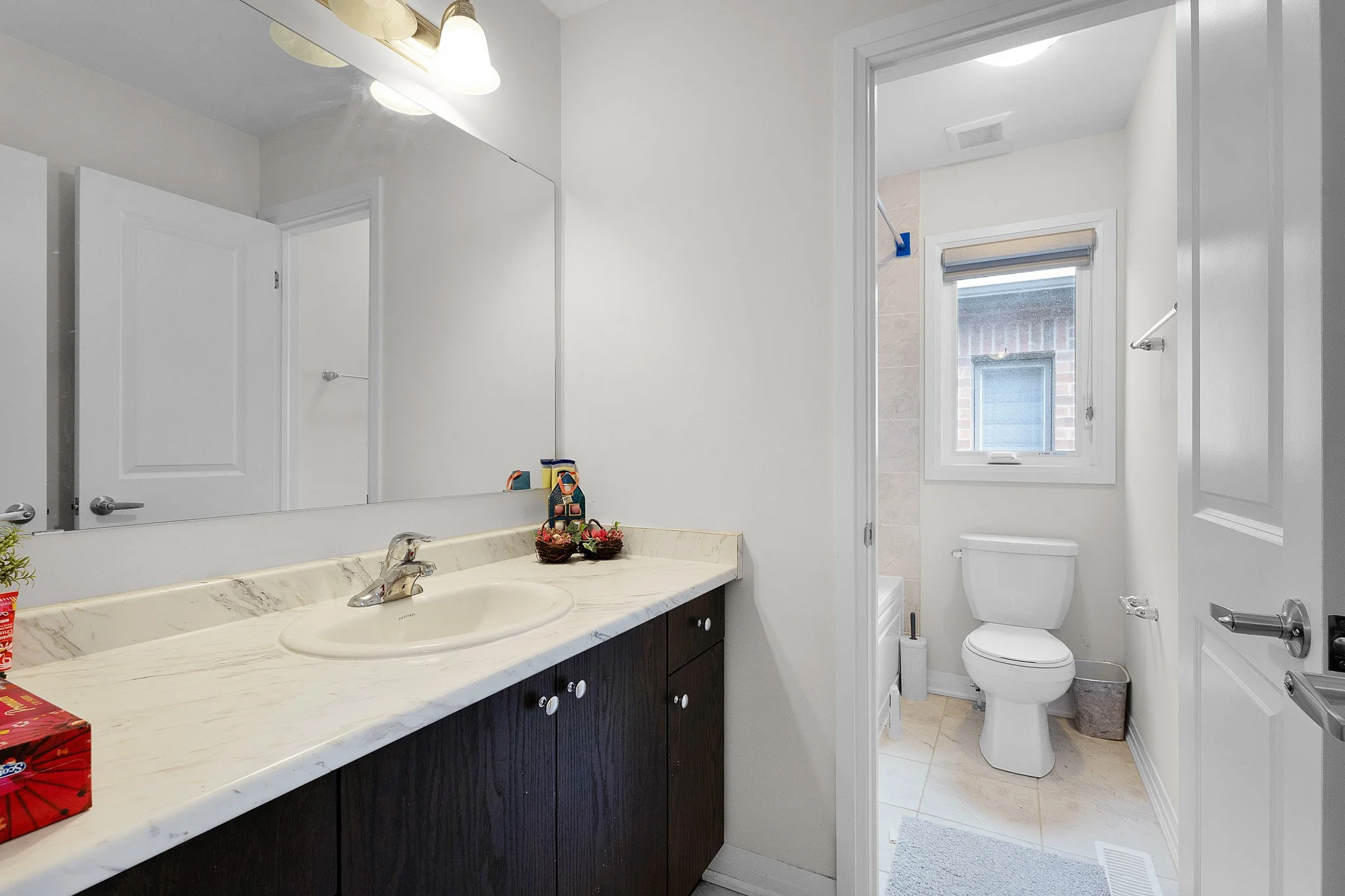 A bathroom with a double sink vanity with a dark wood cabinet and marble countertop, a large mirror, and a small decorative basket of flowers. The room has white walls and a small window above the toilet, which is visible through an open door. A bath