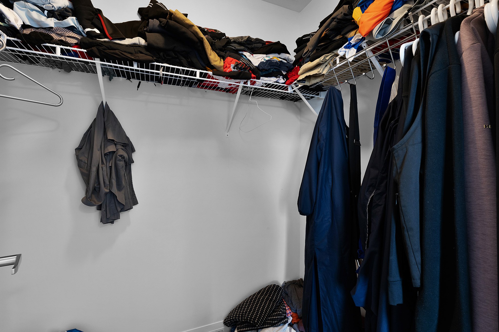 A small walk-in closet with a white wire shelf on the upper wall filled with folded clothes and jackets. Hangers and clothing items hang from hooks, with some clothes stacked or draped. The closet has a white wall and gray carpeted floor.