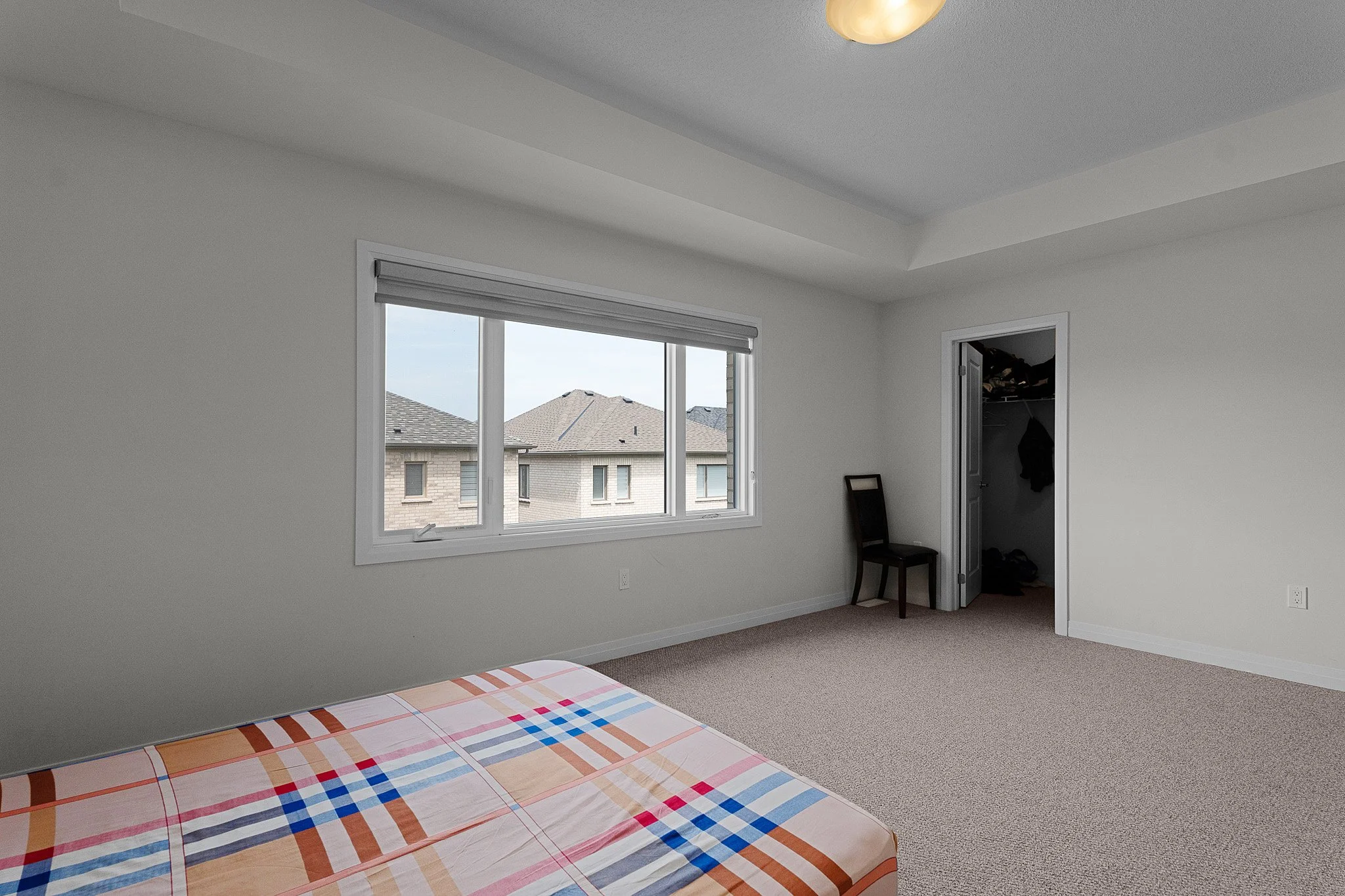 Empty bedroom with a window showing neighboring rooftops, a closet, and a bed with a colorful checkered bedsheet.