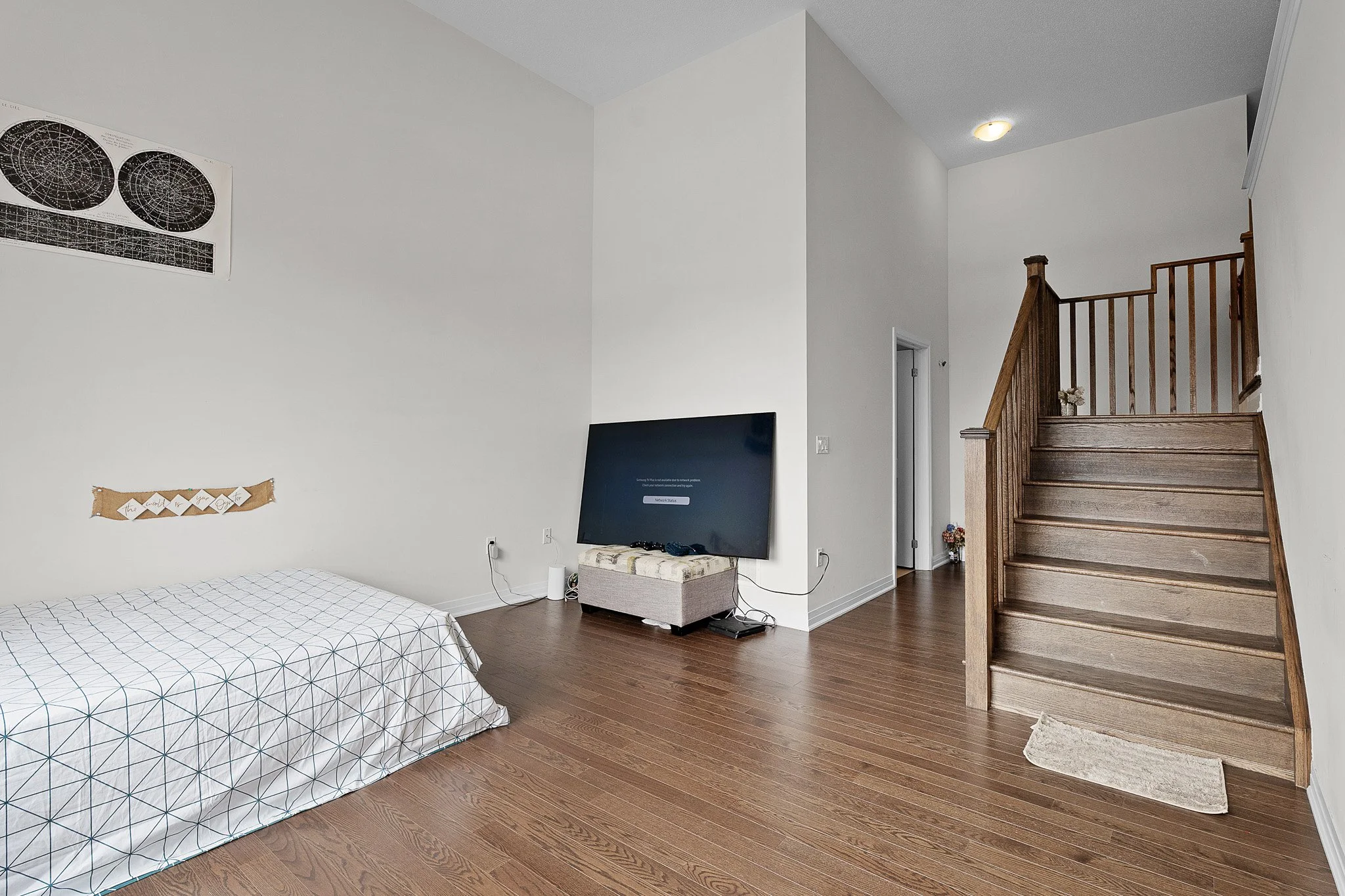 Living room with hardwood floors, a wall-mounted flat screen TV, a bed with geometrically patterned bedding, a small bench under the TV, a staircase with wooden railing, a lighted ceiling fixture, and a wall with polar star and constellation posters.