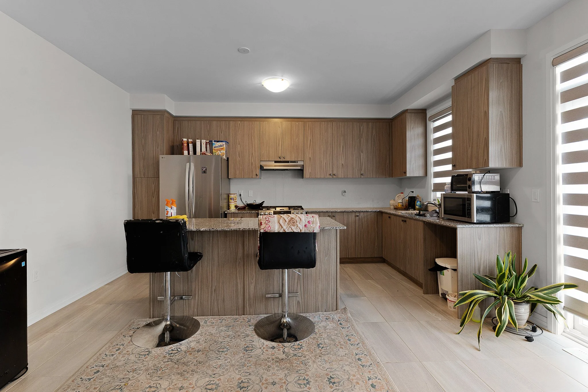 Modern kitchen with wooden cabinets, granite countertops, and stainless steel appliances, including refrigerator and microwave. There is a breakfast bar with two black chairs, a rug on the light wood floor, and a large window with striped blinds. A p