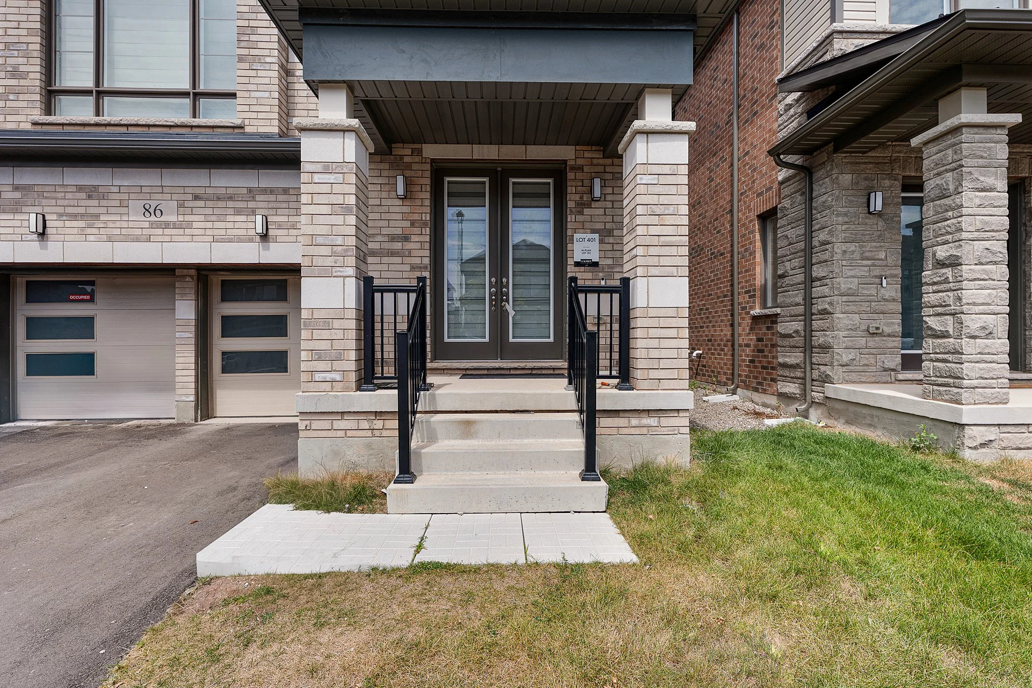Front entrance of a modern brick townhouse with stairs, black railing, and a glass double door. There is a small patch of grass and sidewalk in front, and neighboring buildings with brick and stone facades.