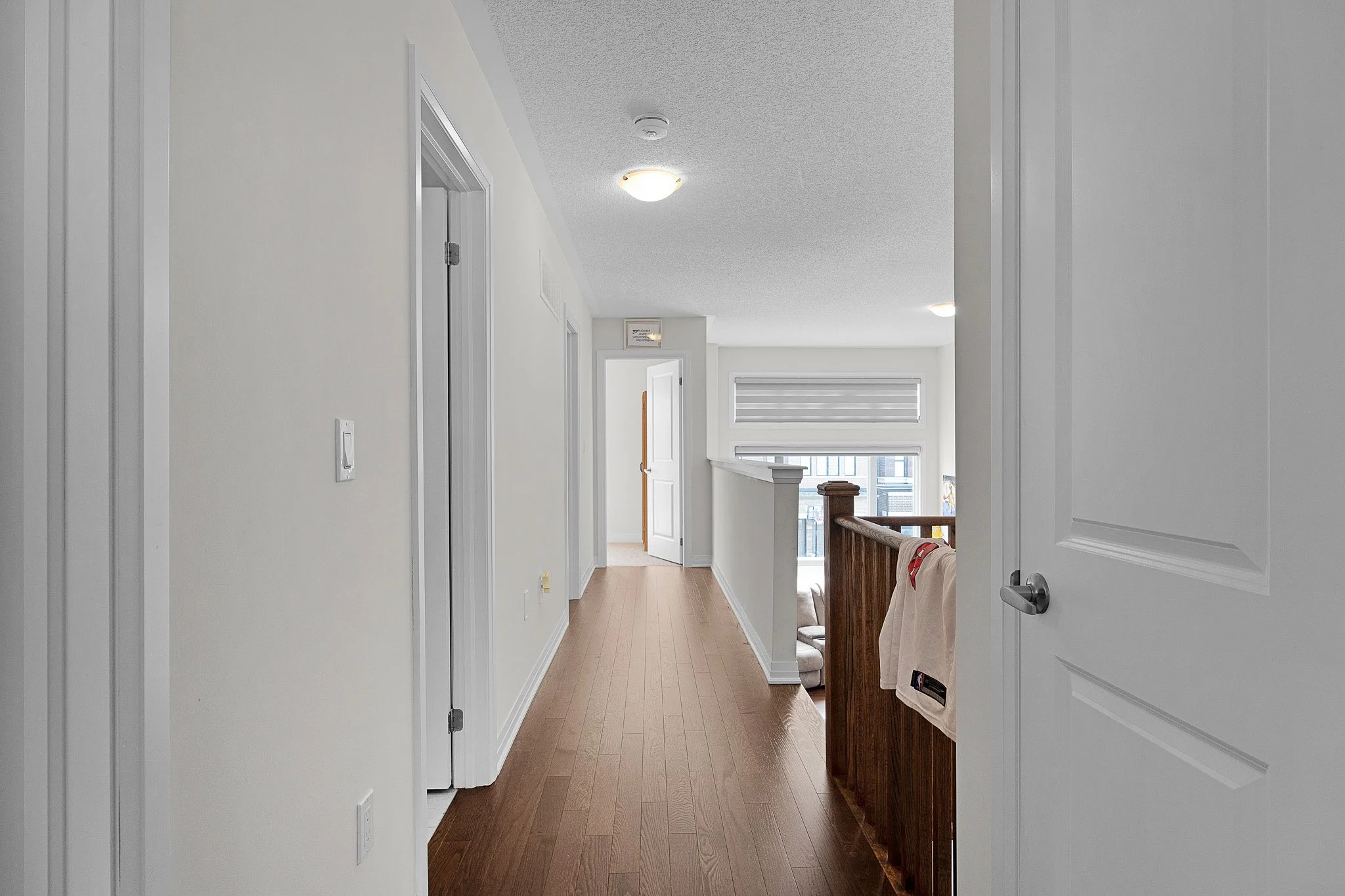View of a bright hallway with white walls and wooden flooring, leading to a balcony with a glass door and window.