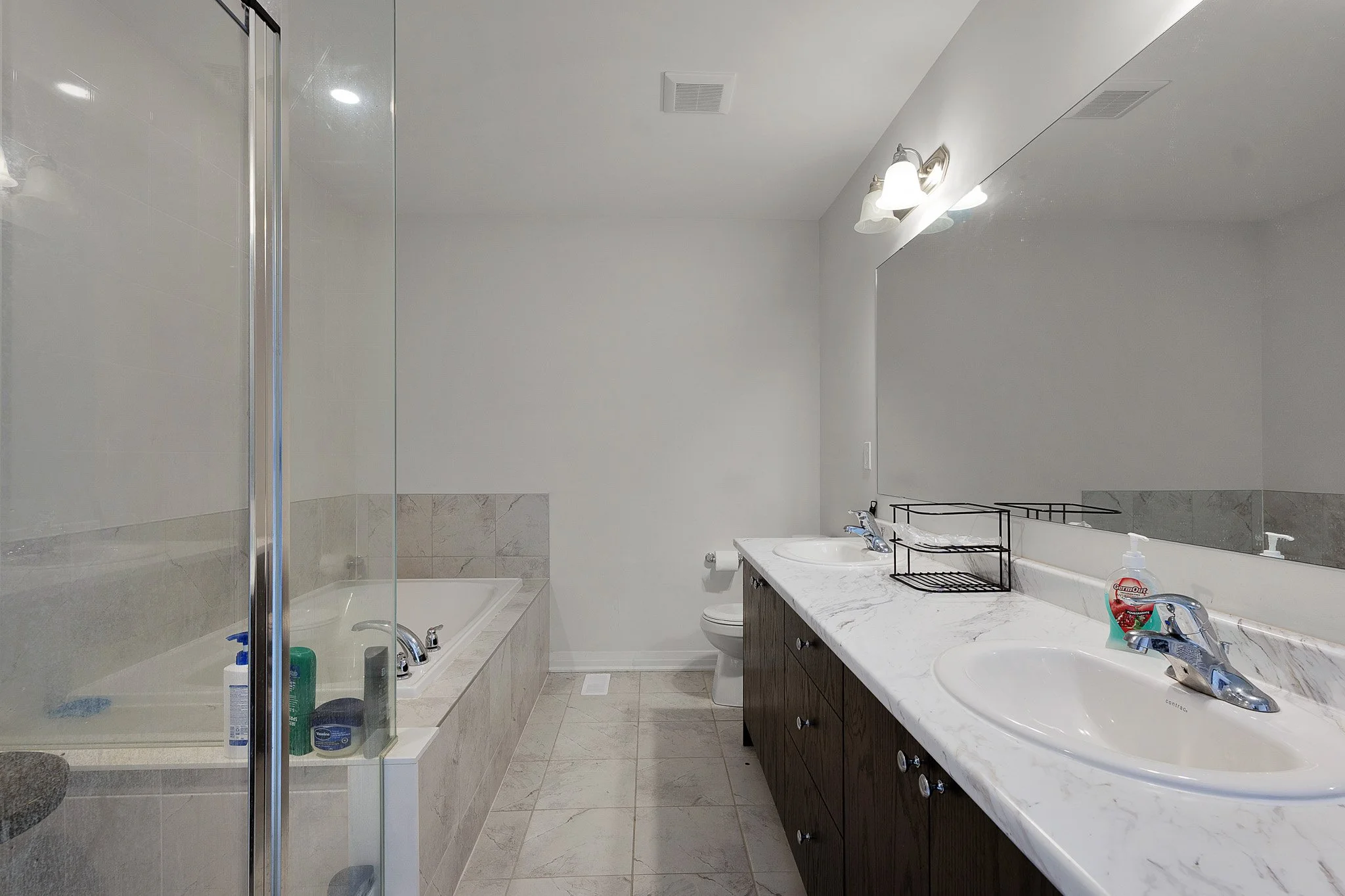 Bathroom featuring a shower area with glass door, a bathtub, a toilet, a sink with a marble countertop, and a large mirror above the sink. The room has white walls, tiled flooring, and built-in lighting fixtures.