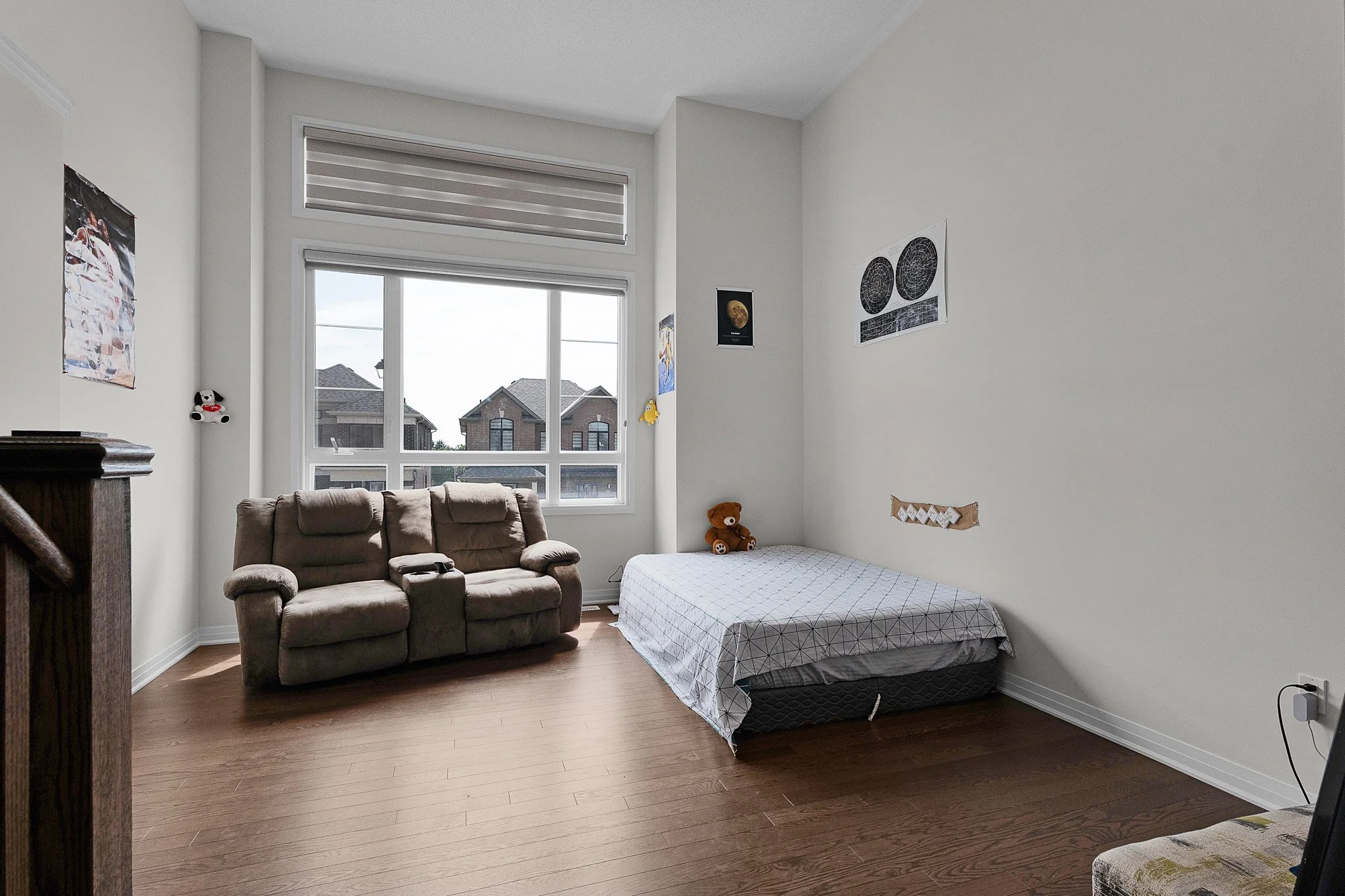 A bedroom with a large window, brown couch, and a single bed with a patterned bedspread. The wall has posters, a small stuffed dog, and a decorative fabric piece. The floor is wooden.