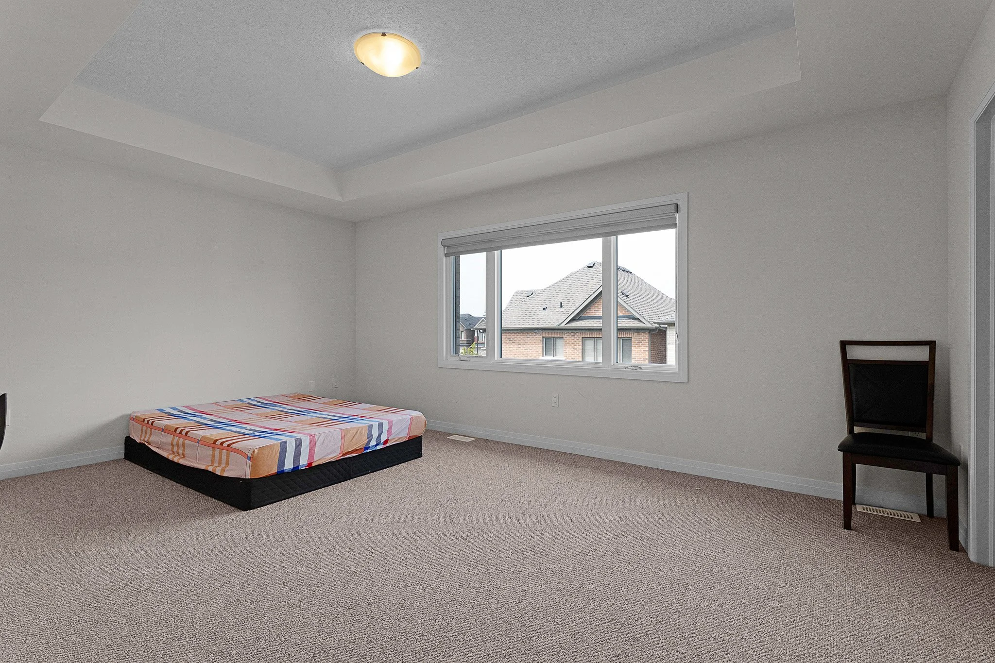 A sparsely furnished bedroom with a window showing a neighboring house. There is a mattress with a colorful striped sheet on a black bed frame and a single chair with a dark frame and cushion. The room has beige carpet and white walls, with a ceiling