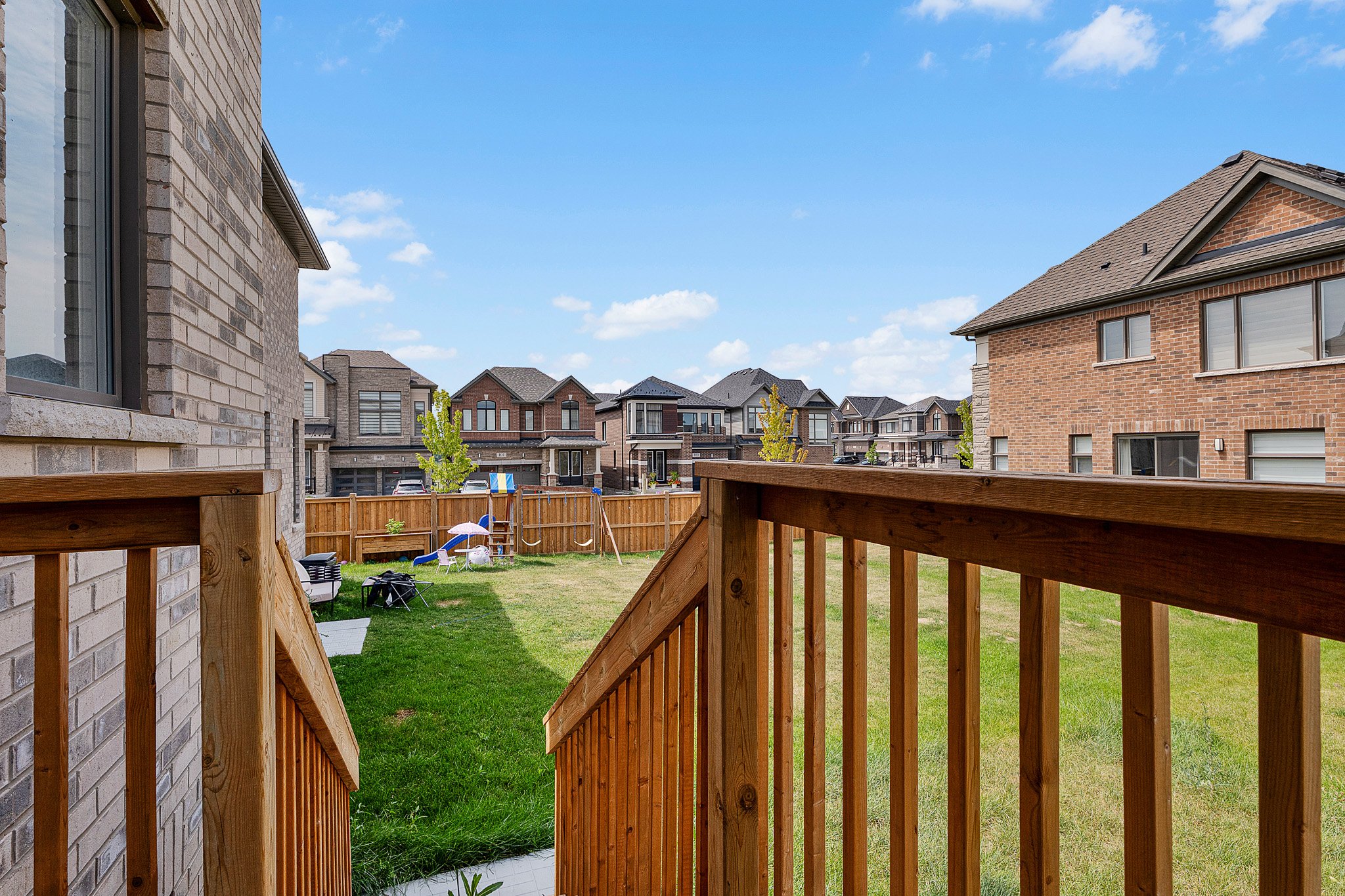 View from a wooden deck looking over a backyard with a grass lawn, fenced yard, and adjacent houses in a suburban neighborhood under a partly cloudy sky.