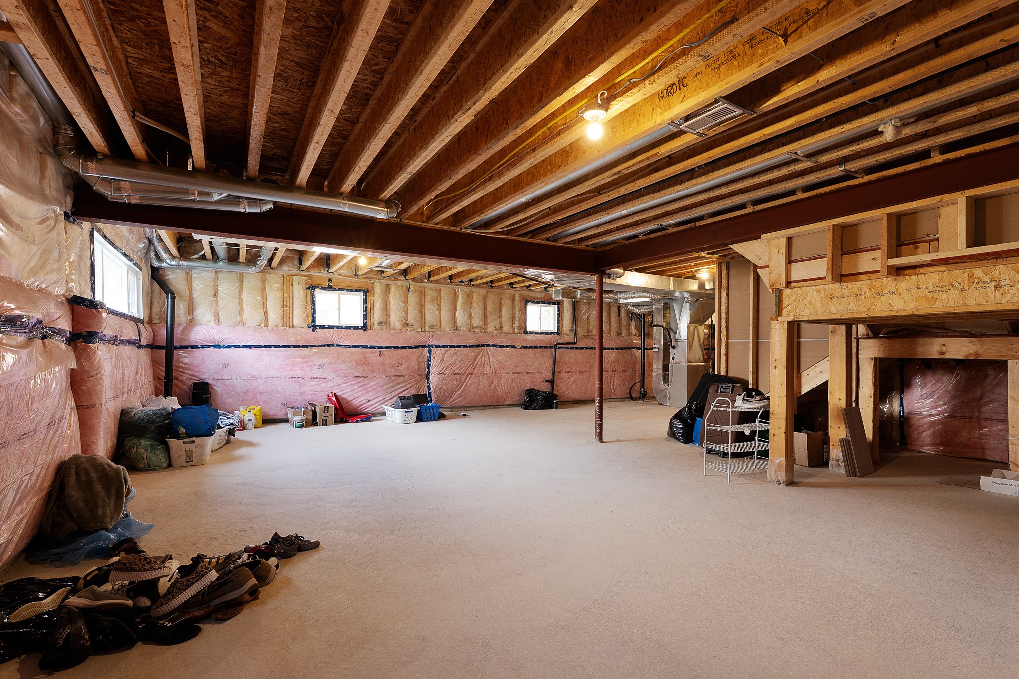 Unfinished basement with exposed wooden ceiling joists, ductwork, and insulation on one wall. Carpeted floor with shoes, storage bins, and tools. Windows and construction materials are visible.