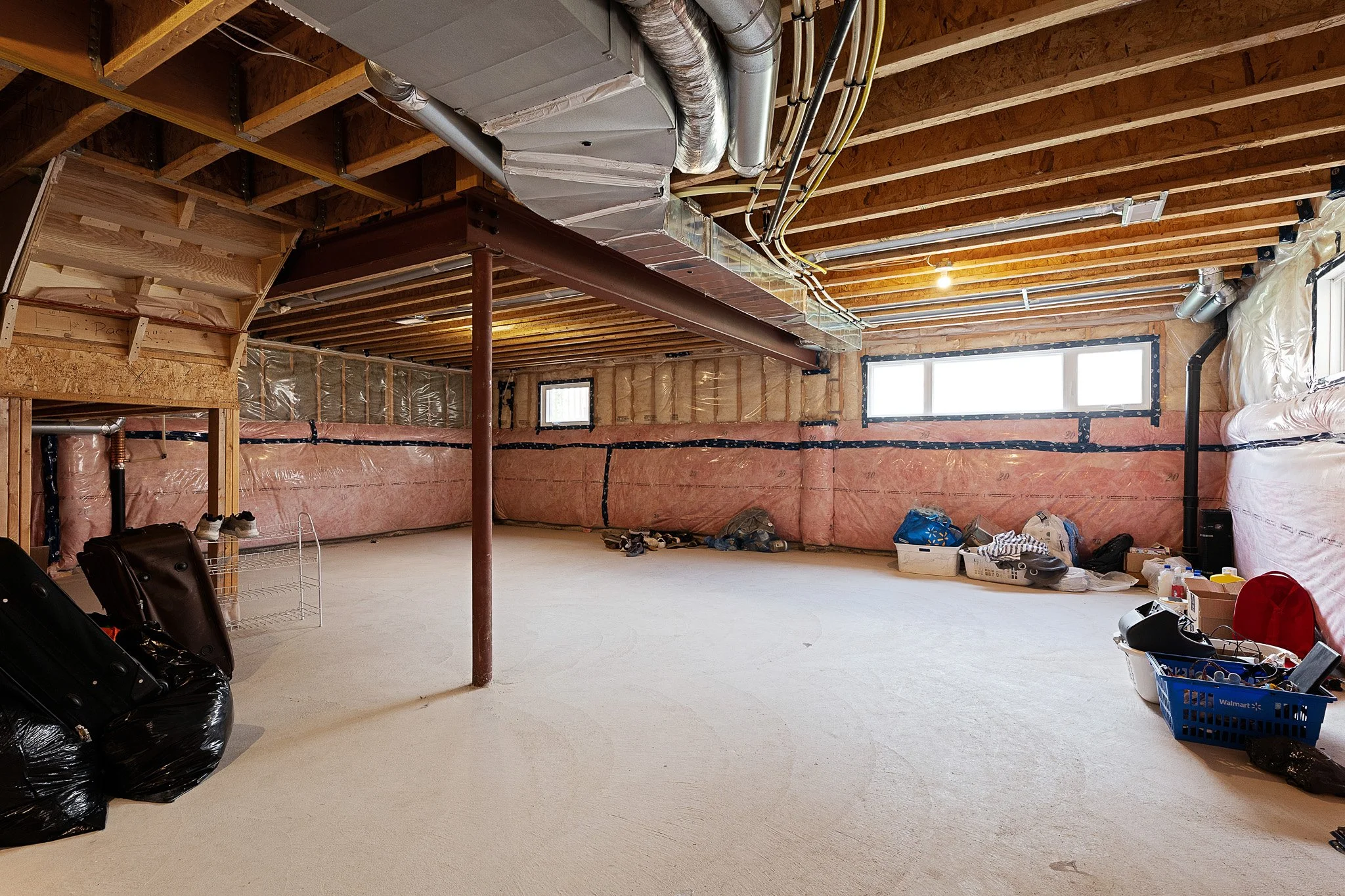 An unfinished basement with exposed ceiling beams, ductwork, and insulation along the walls, a window on the right, and clutter on the floor including bags, boxes, and shoes.