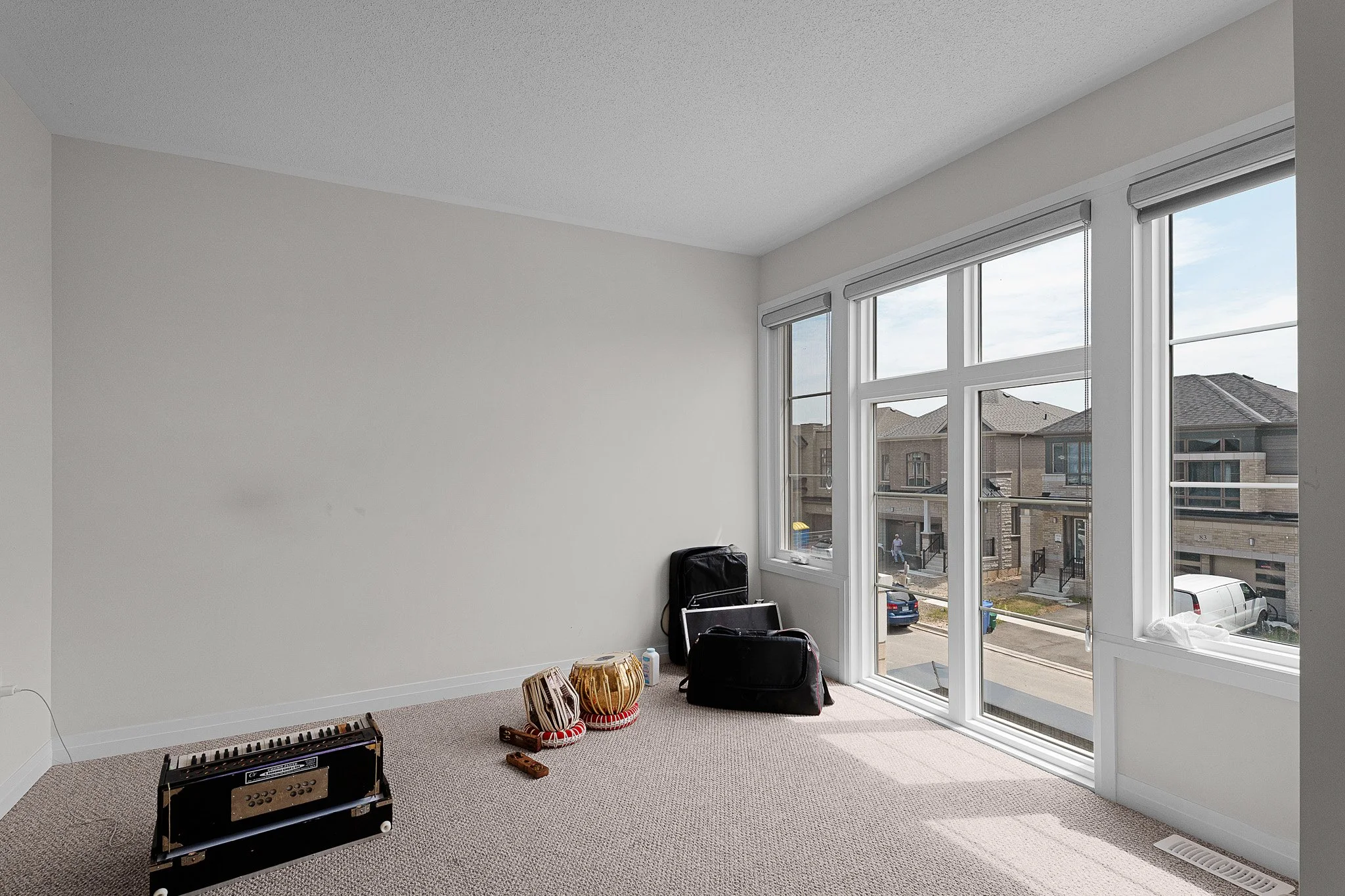 Empty room with large windows and a view of neighboring houses. Room contains a small black amplifier, baskets, and some other miscellaneous items on the floor, with wall and ceiling painted white.