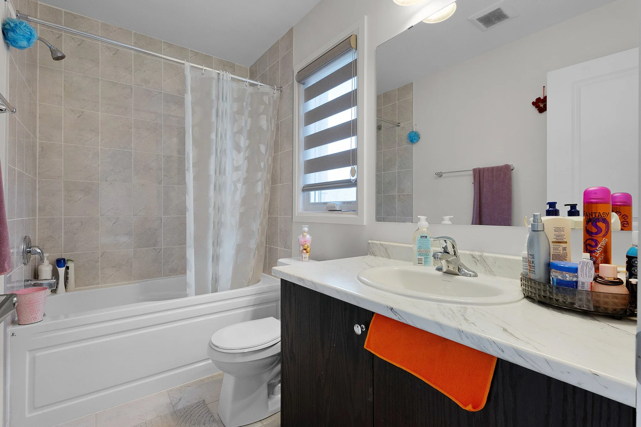A bathroom with a bathtub, shower, toilet, and a marble countertop with various toiletries. A window with a striped roller shade above the sink. Towel and decorative items on the walls and countertop.