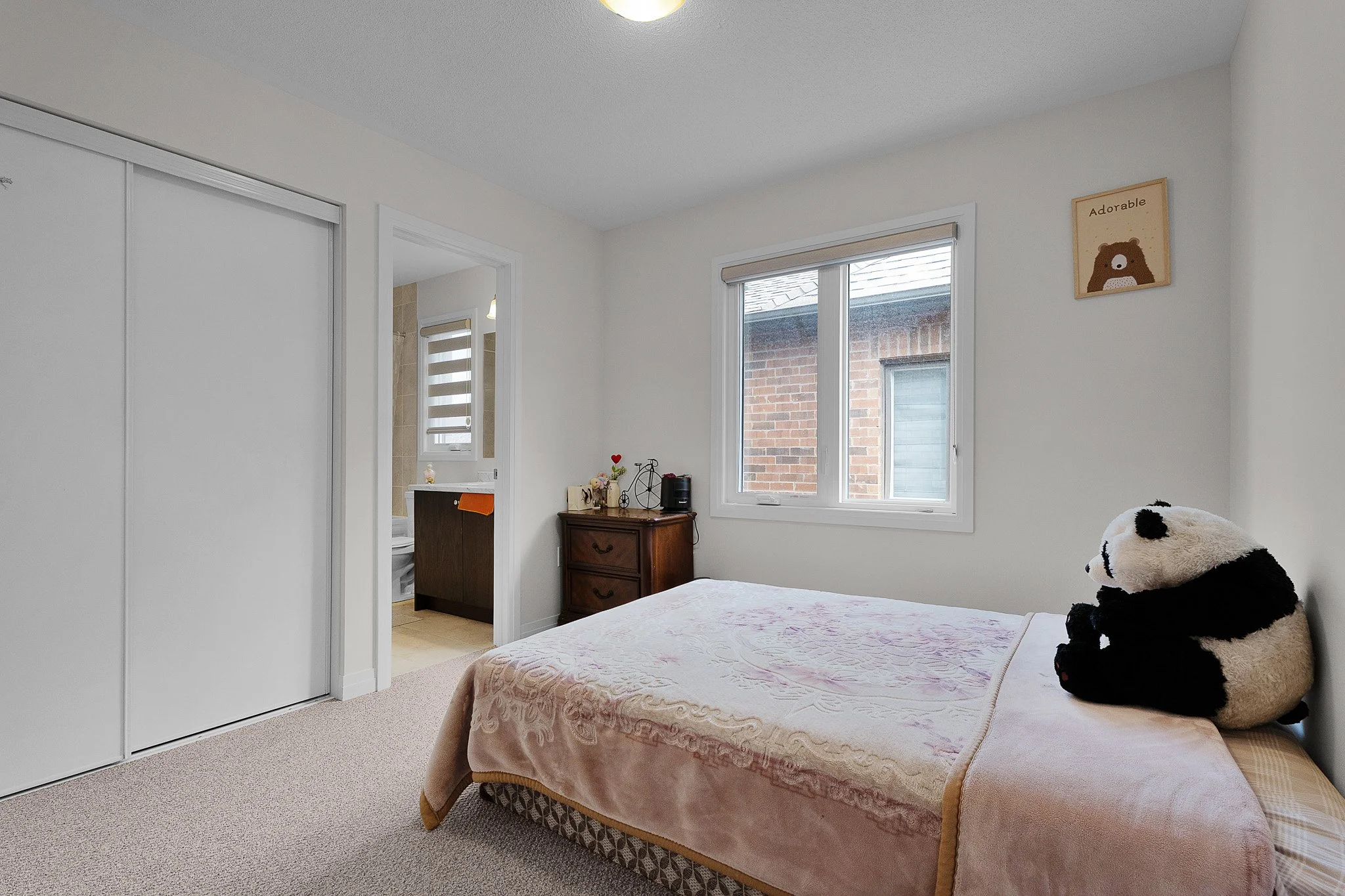 A bedroom with a bed covered in a light pink quilt and a stuffed panda bear on it. There is a small wooden dresser with flowers and decorative items next to the bed. Two windows with white frames and blinds, showing a brick wall outside. A closet wit