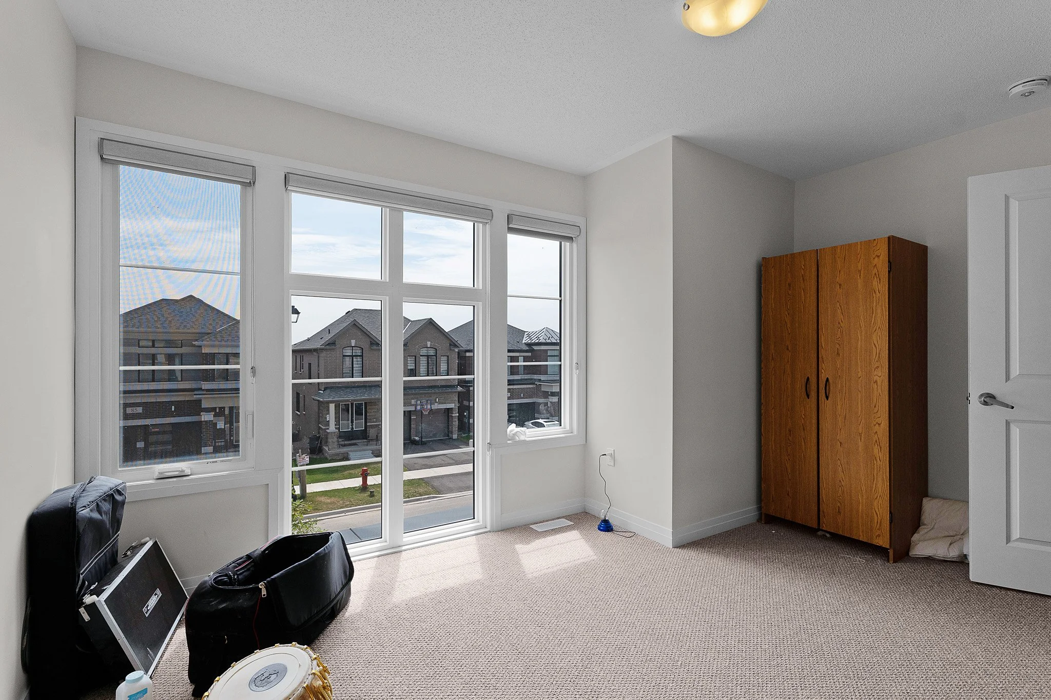 Empty room with large windows showing residential neighborhood, some clutter including a guitar case, a luggage bag, and musical instruments, a wooden cabinet, white walls, and beige carpet flooring.