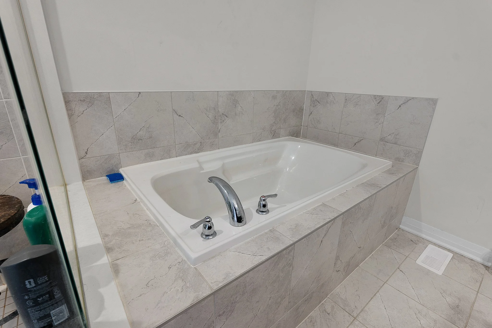 Bathroom with a built-in bathtub surrounded by light gray marble tiles, with a silver faucet and handles, located in a tiled room with a heating vent on the floor.