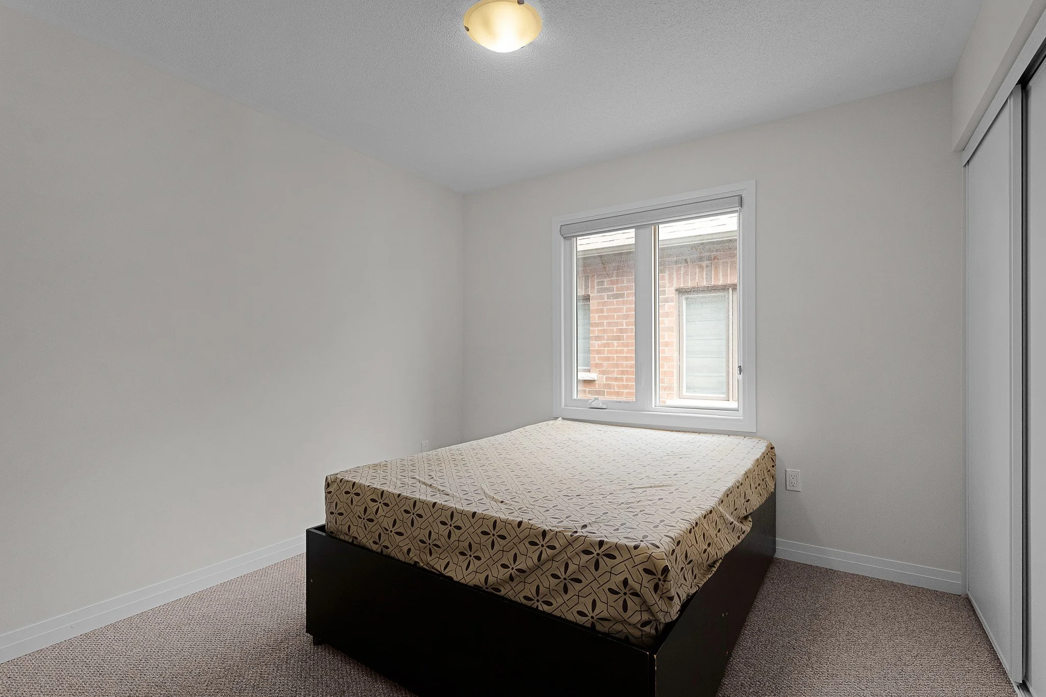 A minimalist bedroom with white walls and a window showing a brick building outside. There is a bed with a brown geometric patterned bedspread and no headboard, positioned near the window. A ceiling light fixture is mounted on the white ceiling.