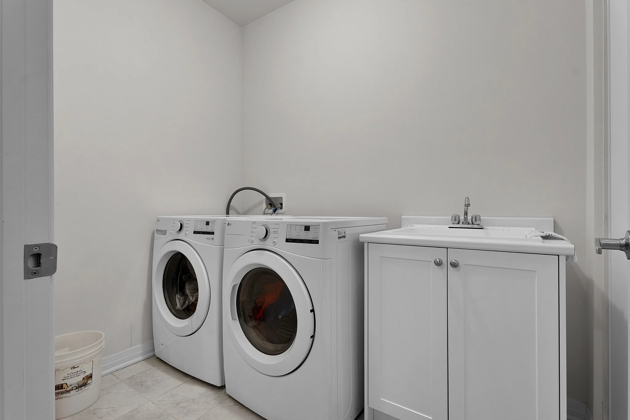 A laundry room with white walls, a white washing machine and dryer side by side, a white cabinet with a sink, a small white trash bin, and an empty wall space.