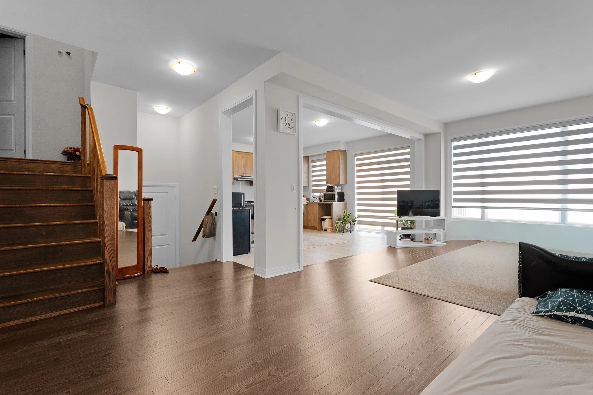 View of a modern living room with wooden flooring, a white couch with pillows, a TV on a white stand, and large windows with striped blinds, with part of a staircase visible on the left.