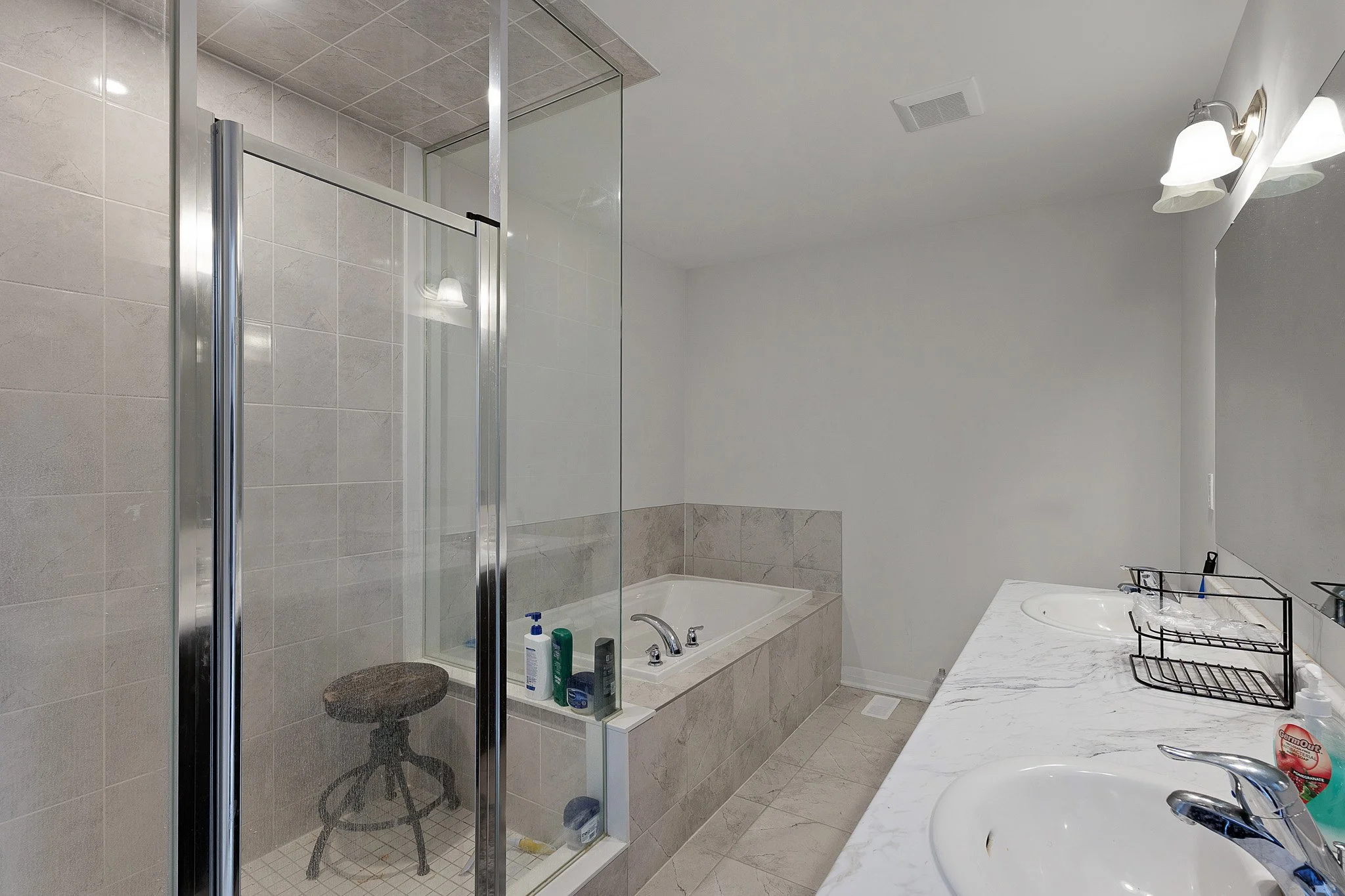 A bathroom with a glass shower enclosure, bathtub, white sink, and marble countertop. The shower has a metal frame and a stool inside. There are toiletries on the bathtub ledge. The wall is tiled with beige tiles, and the ceiling is white with a vent