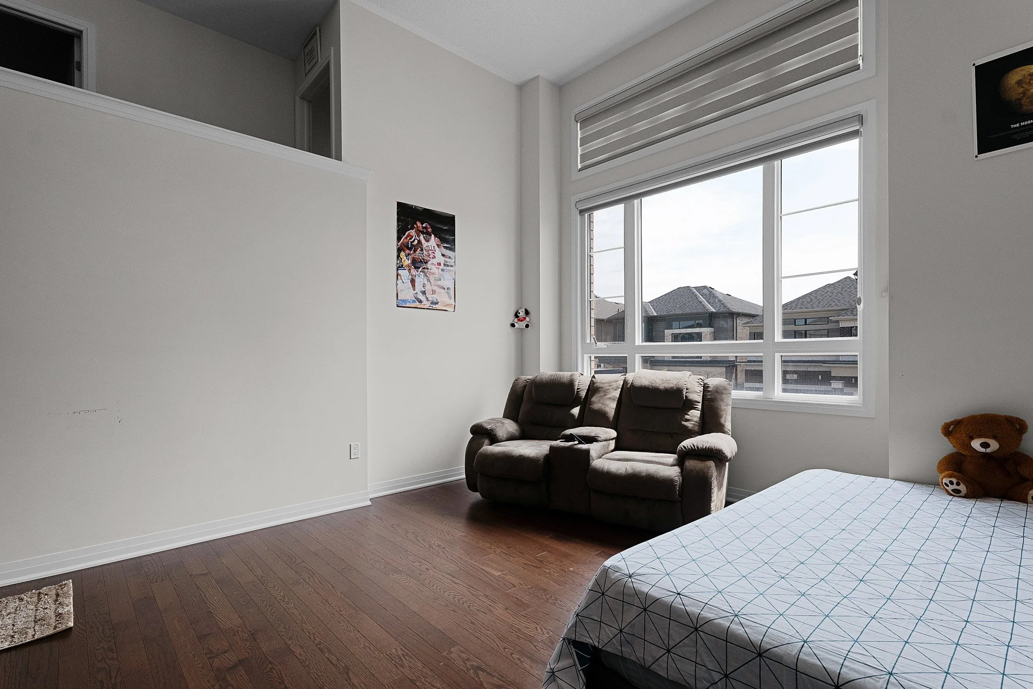 A bedroom with large windows, a brown reclining chair, a teddy bear on the bed, a poster of basketball players on the wall, and hardwood floors.