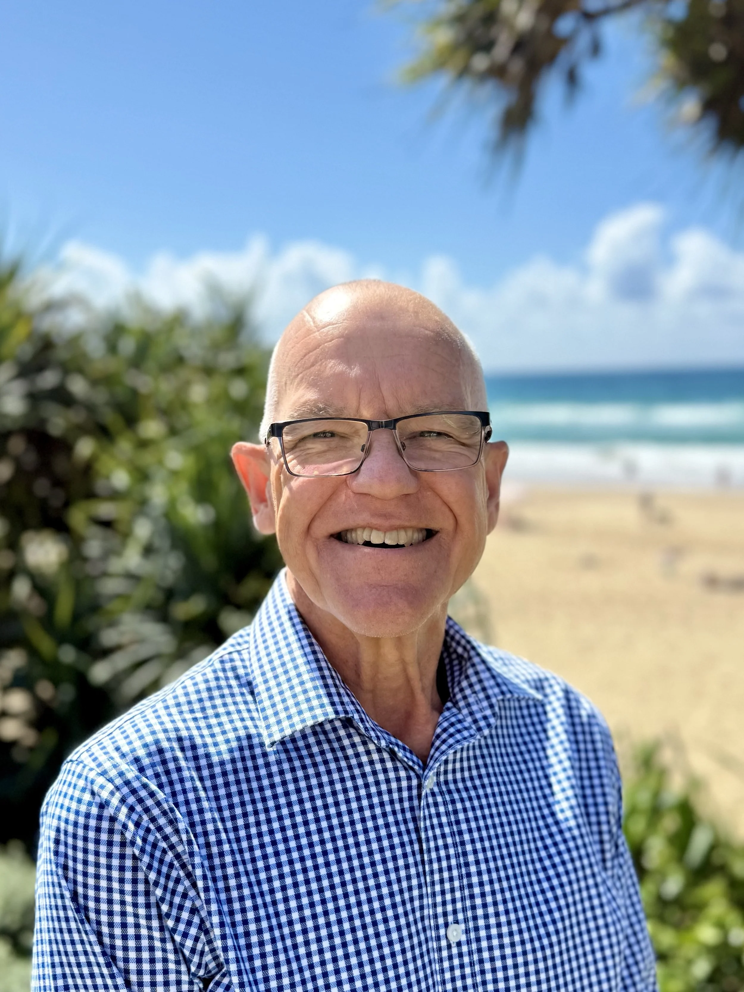 An elderly man with glasses smiling outdoors near the beach with sand, ocean, and sky in the background.