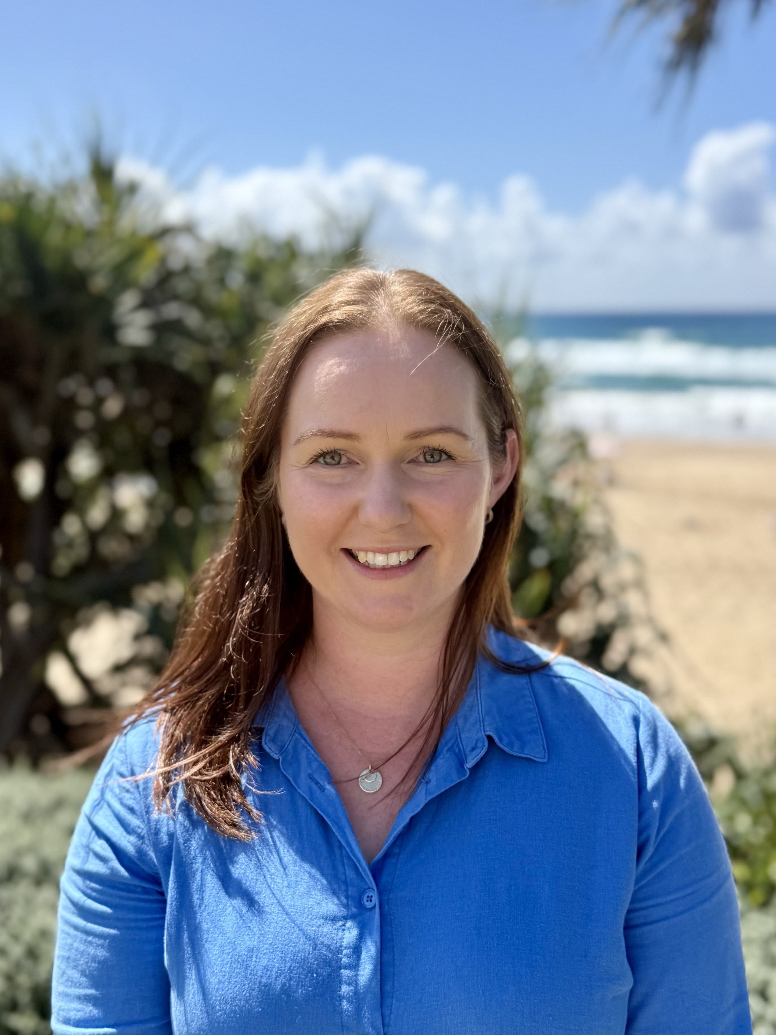 A woman with reddish-brown hair smiling outdoors near the beach with green plants and ocean in the background.