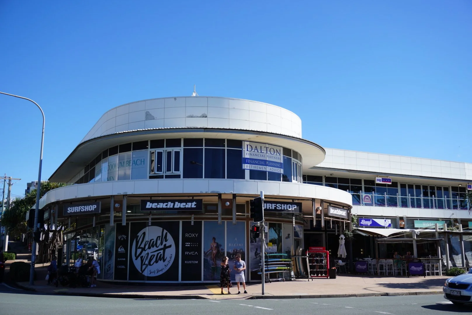 A modern corner building on a busy street with various storefronts, including Surfshop and Beach Beat, featuring surfboards displayed outside. No people are visible walking on the sidewalk.