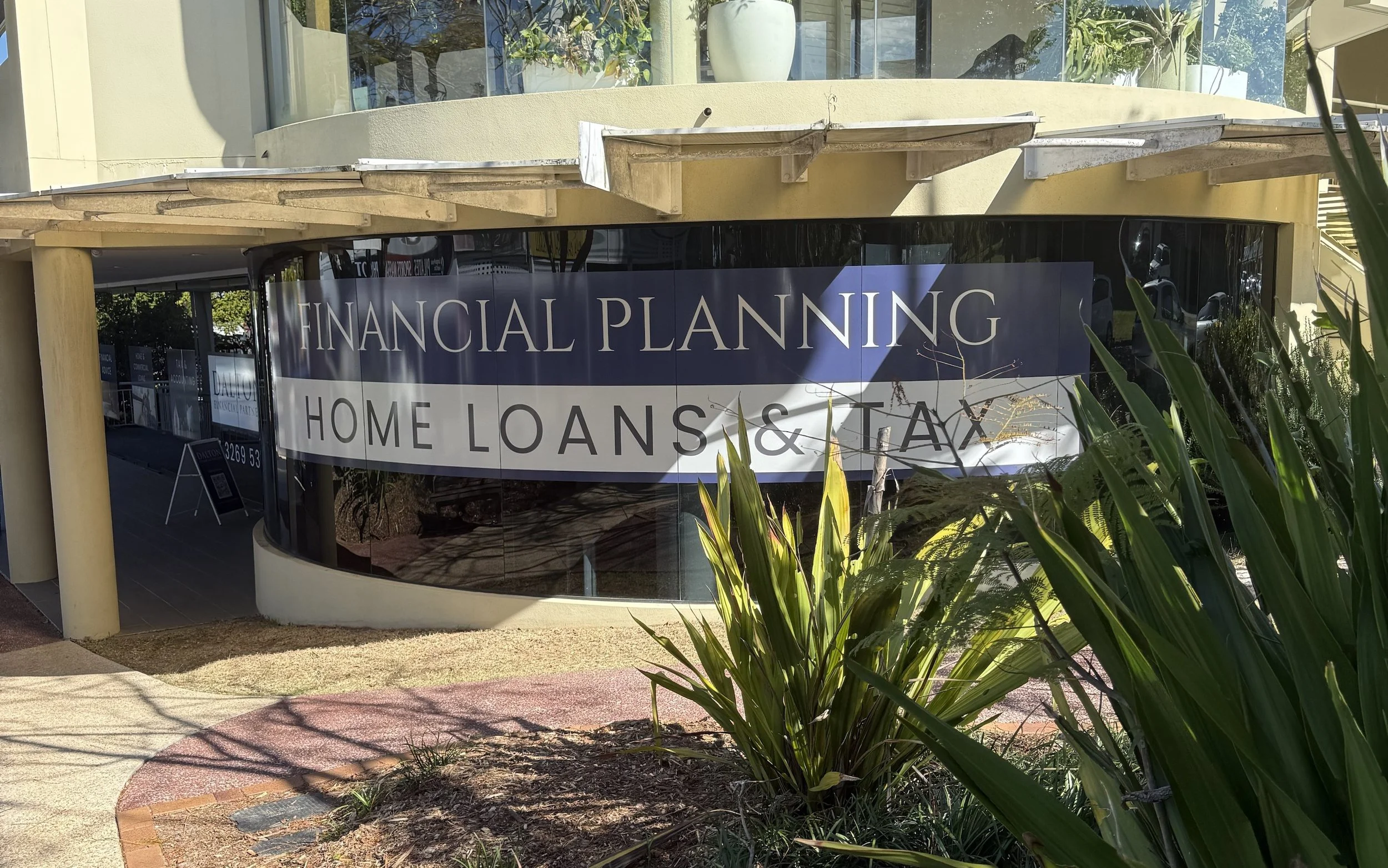A storefront with sign reading 'Financial Planning Home Loans & Tax' reflected in large curved glass windows, surrounding greenery and a sidewalk in front.