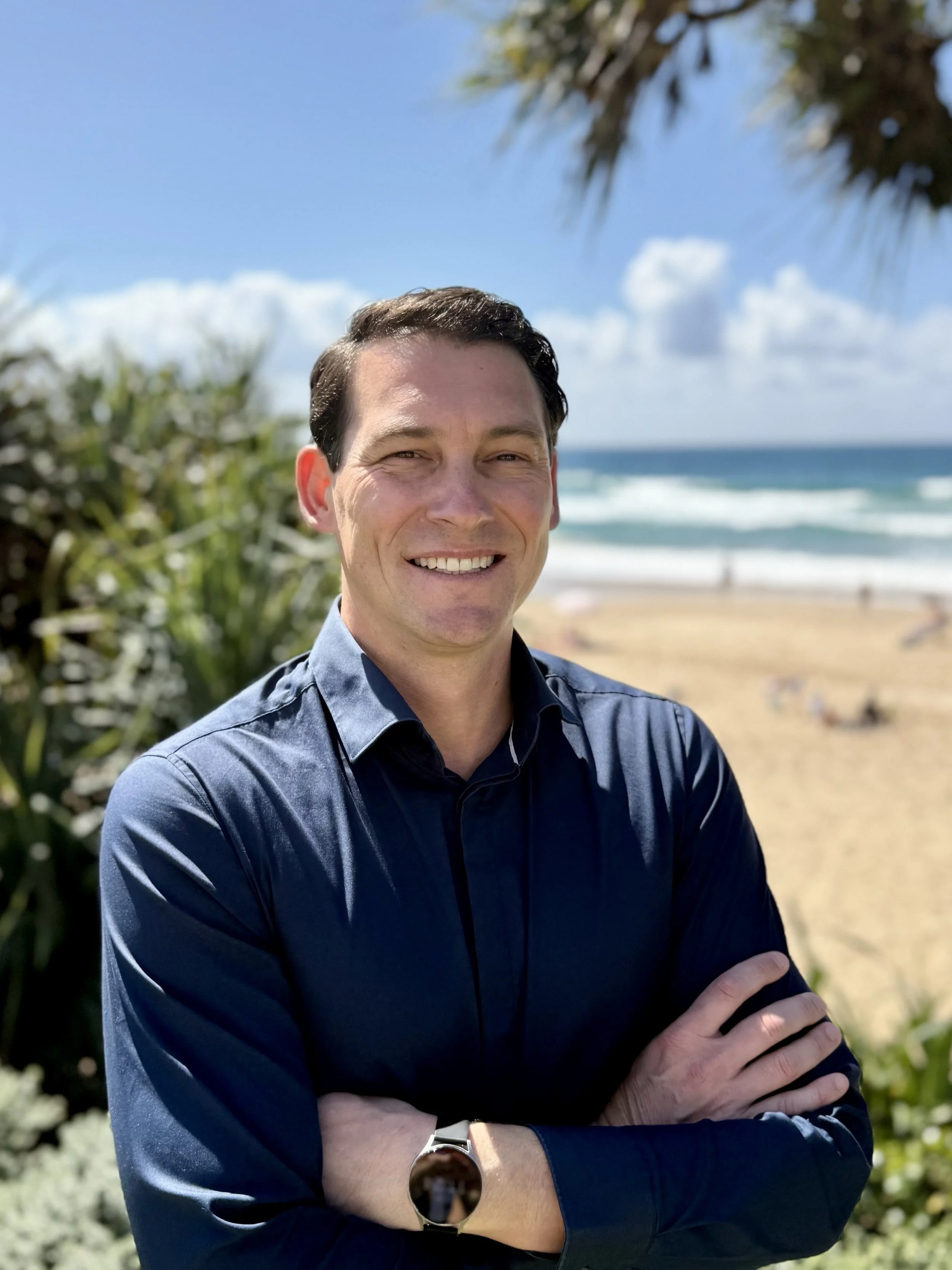 Smiling man in a navy shirt standing outdoors near the beach with ocean waves and green plants in the background.