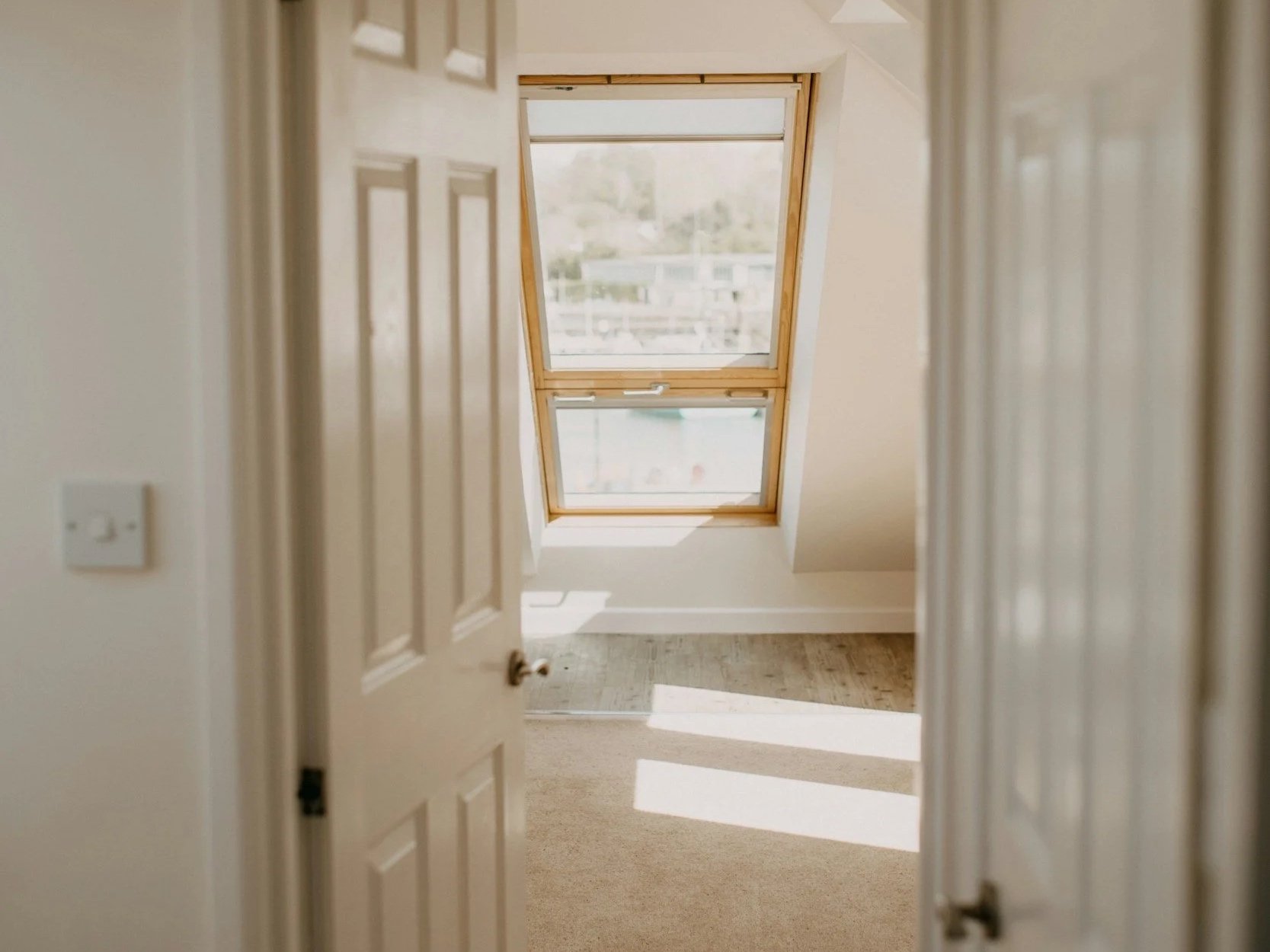 View through an open white door into an empty attic room, with sunlight from a skylight falling across the carpet.