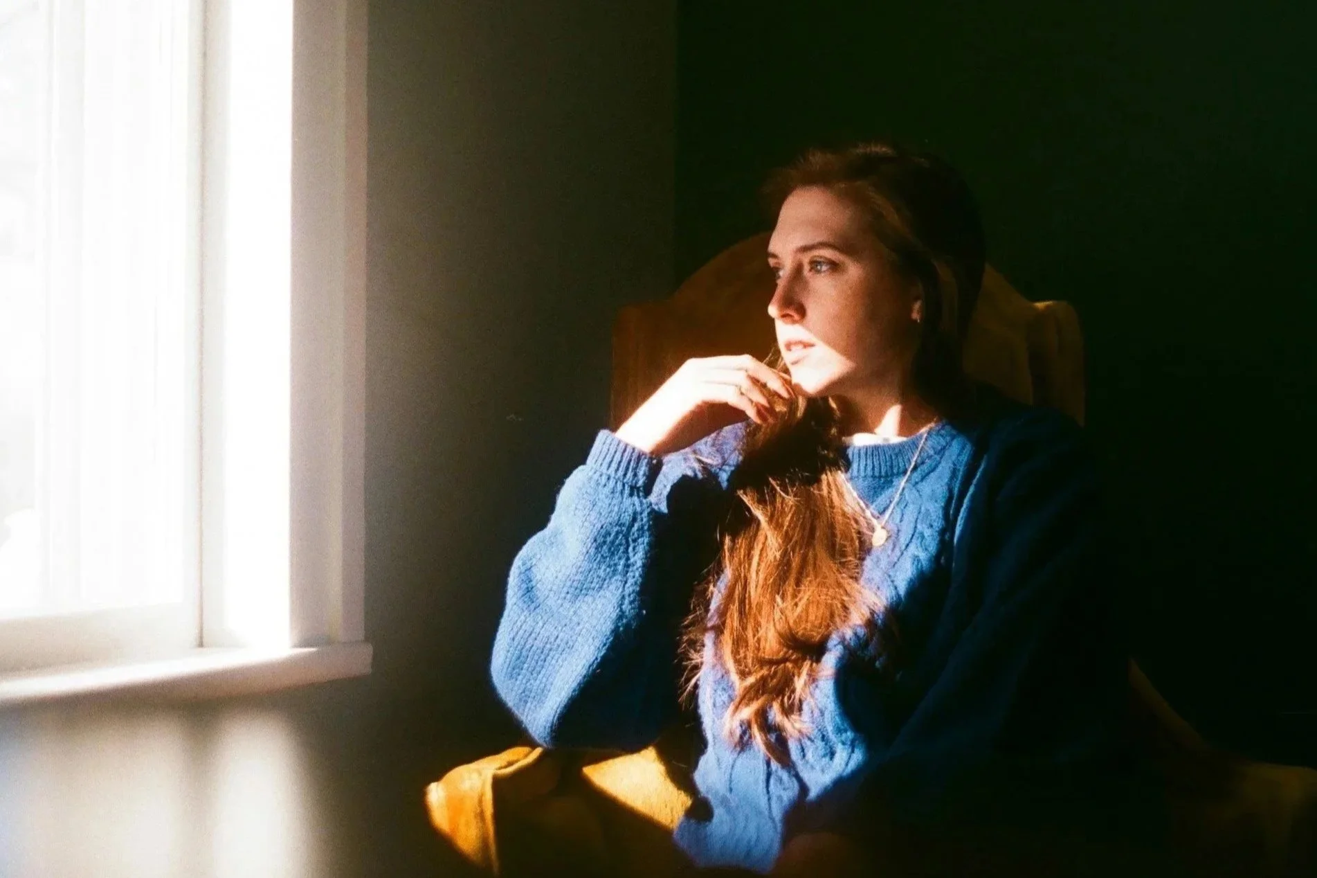 A woman in a blue sweater sitting in a chair near a window, looking outside in soft natural light with a thoughtful expression.