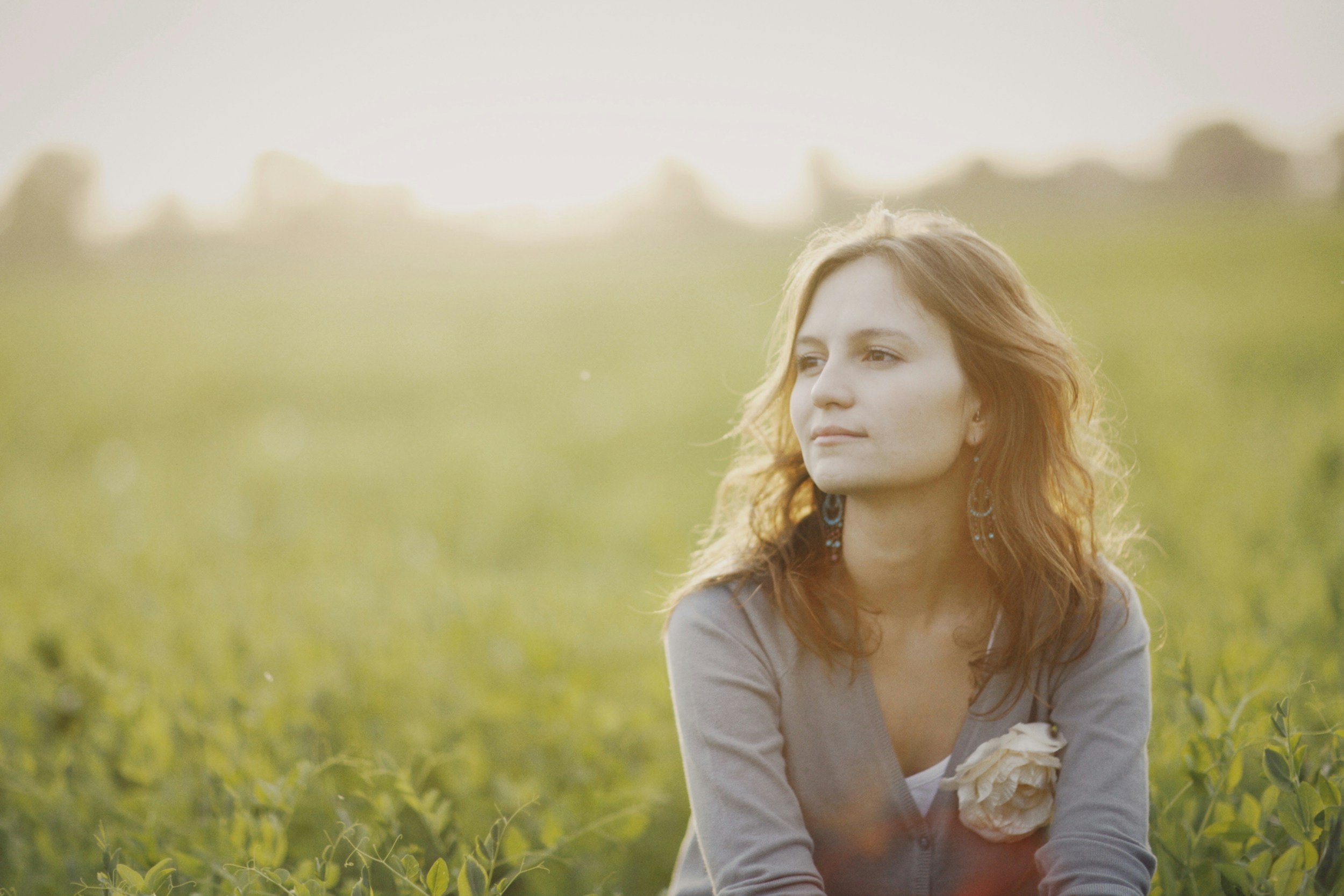 A woman with wavy hair sits in a sunlit green field, gazing quietly to the side with a calm and open expression.