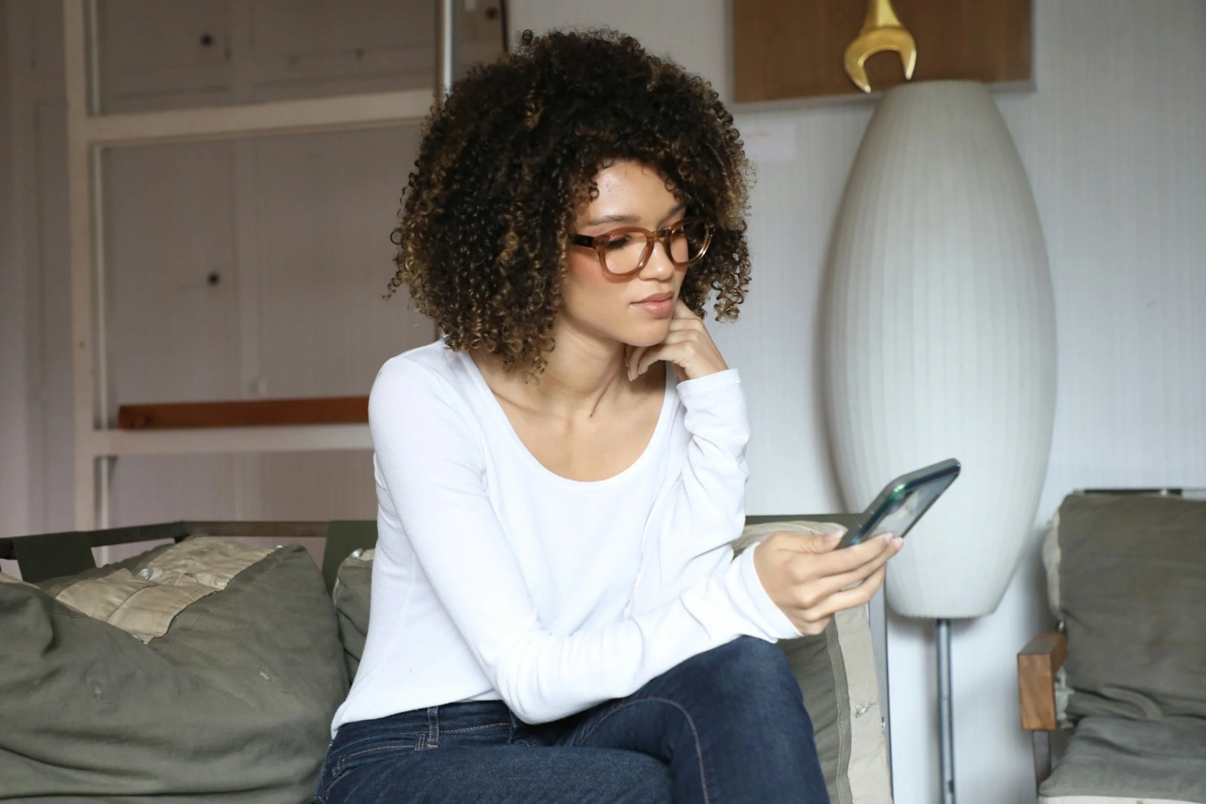 A woman with curly hair and glasses sits on a couch, chin resting on her hand, looking thoughtfully at her phone.