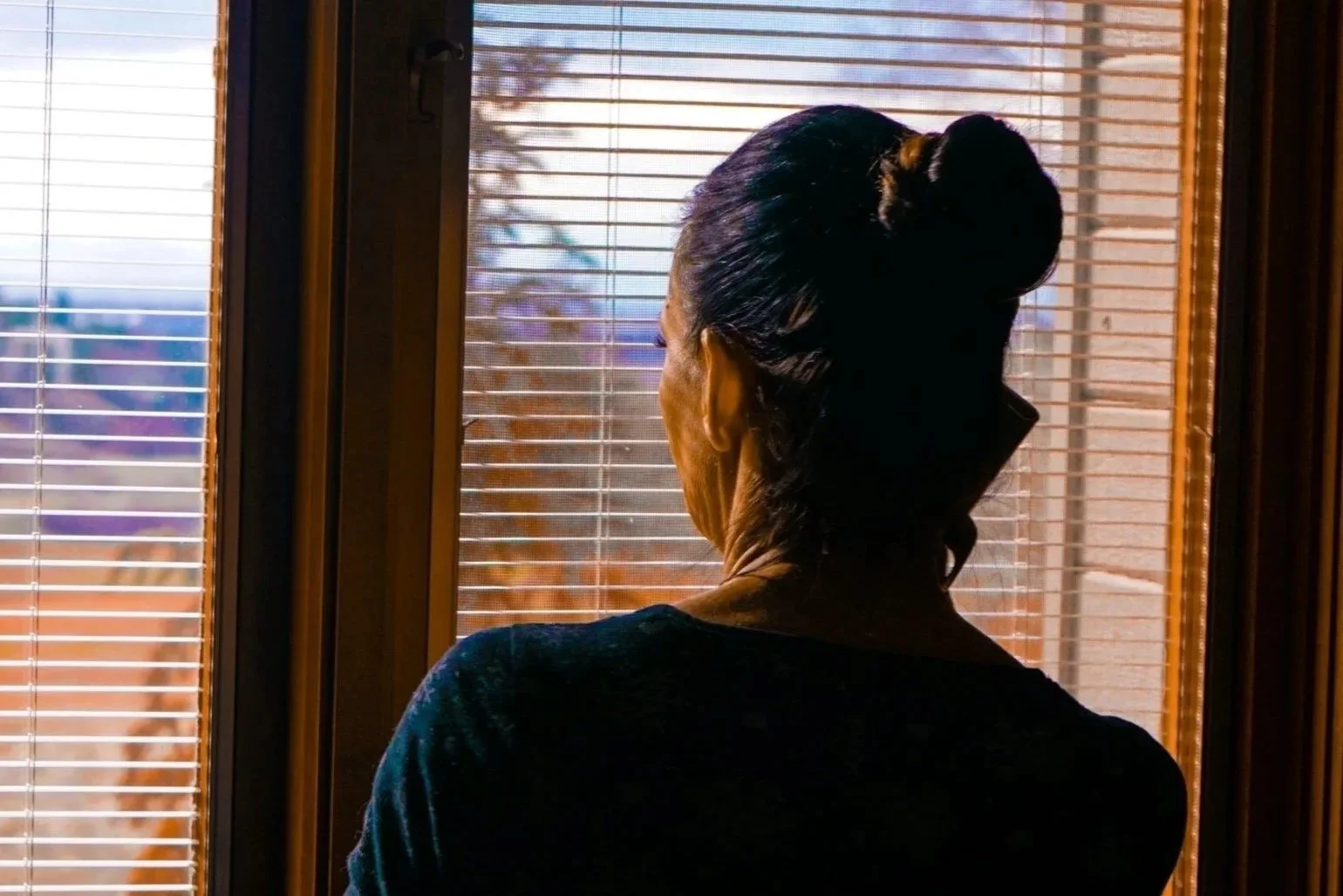 Woman standing by a window in soft light, looking outside in a quiet interior setting.
